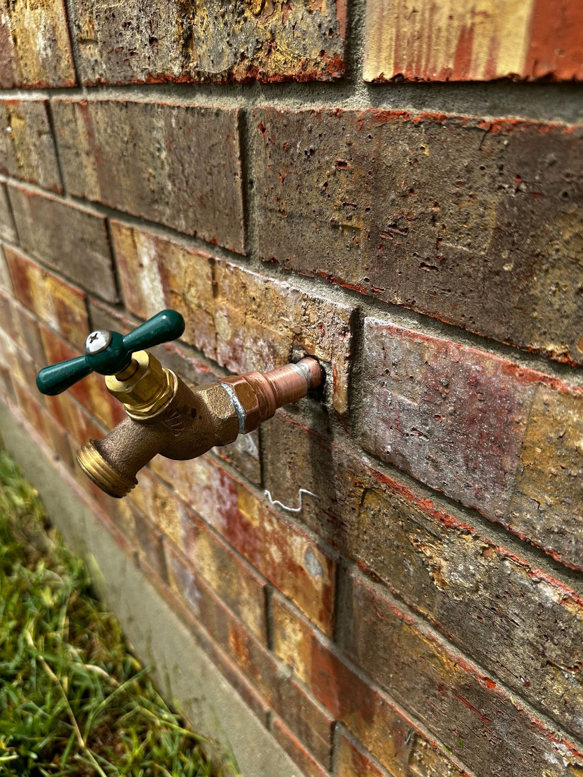 A faucet with a green handle is attached to a brick wall.