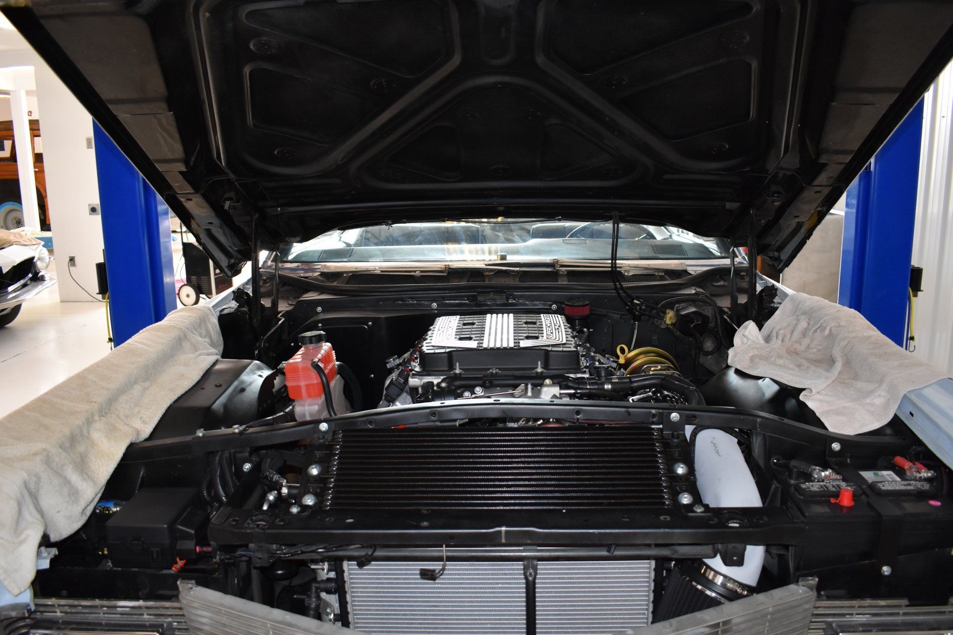 Car engine bay with hood open in a garage, showing the front grille and engine components.