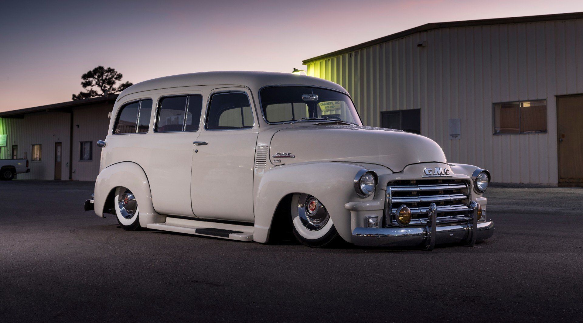 Low white vintage van parked on pavement beside an industrial building at dusk