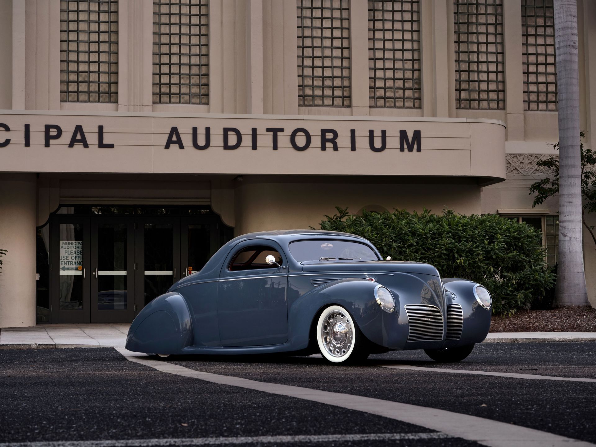 Blue vintage custom car parked outside a municipal auditorium.