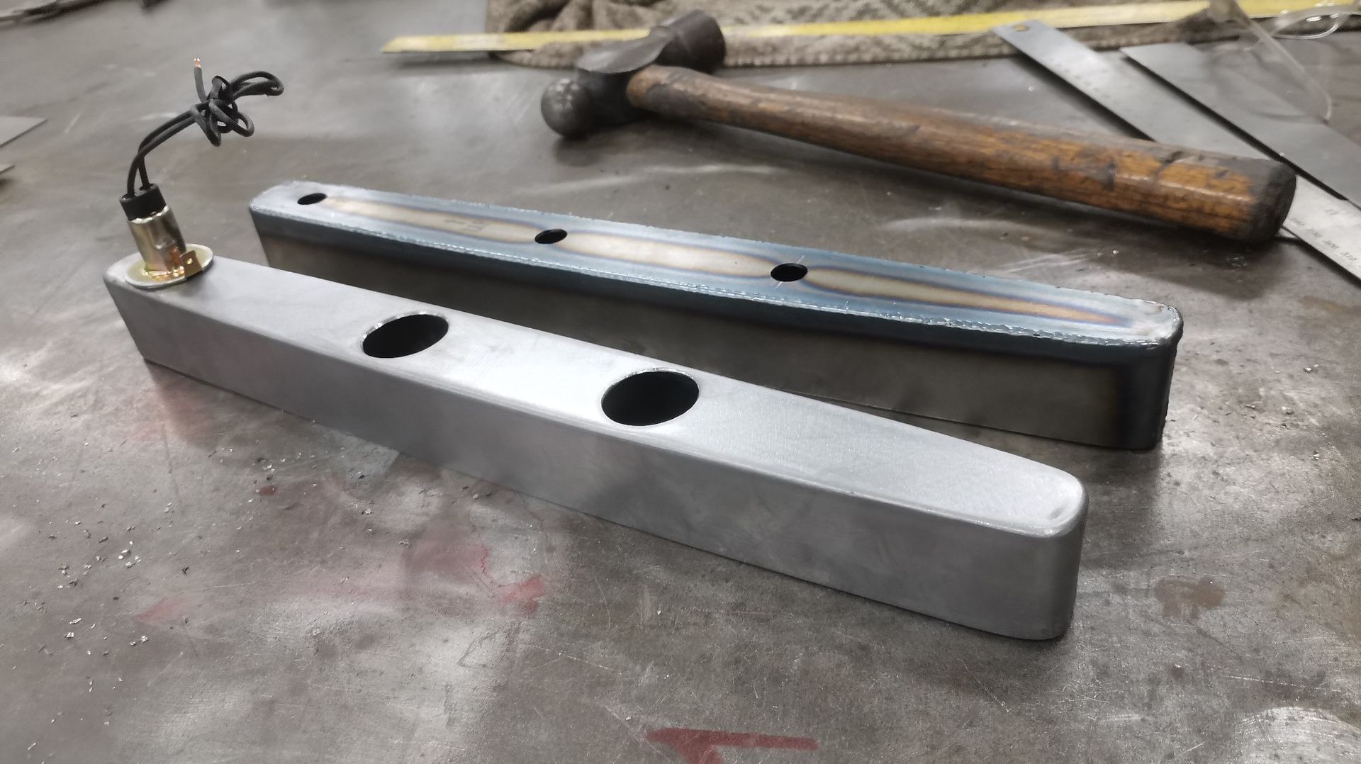 Two metal bars on a workbench, one silver with holes and one black, with hammers in the background