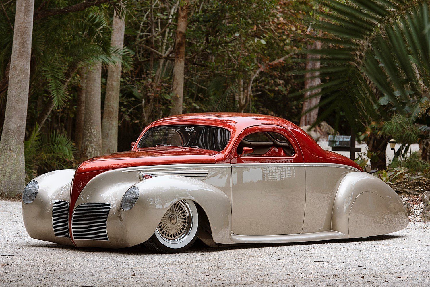 Custom red-and-cream vintage hot rod parked on a gravel path in a wooded garden