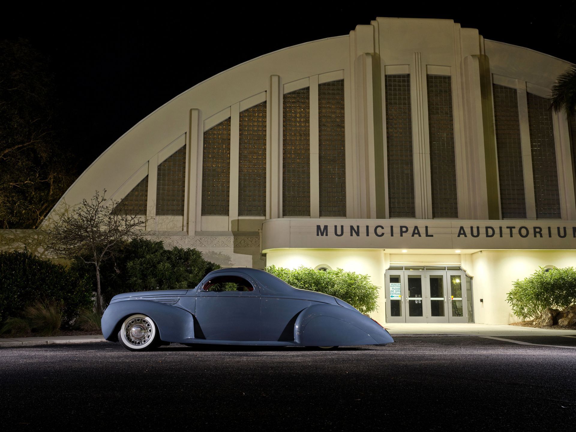 Blue vintage car parked outside a lit municipal auditorium at night