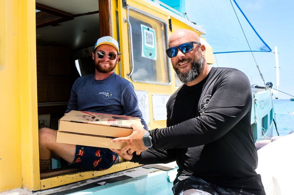 Two men are standing next to each other on a boat holding pizza boxes.