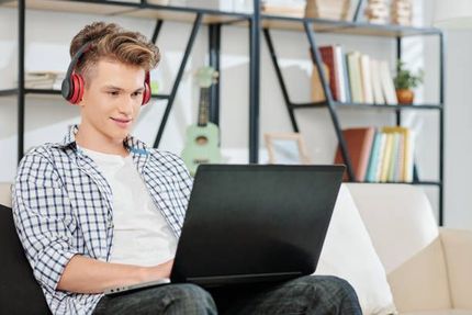 Woman with blonde hair sits at a wooden table in front of a laptop, smiling. White wall background.