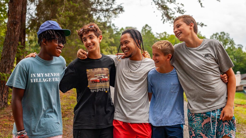 Group of five smiling teens outdoors, two girls flanking the central male figure, two others hugging him.