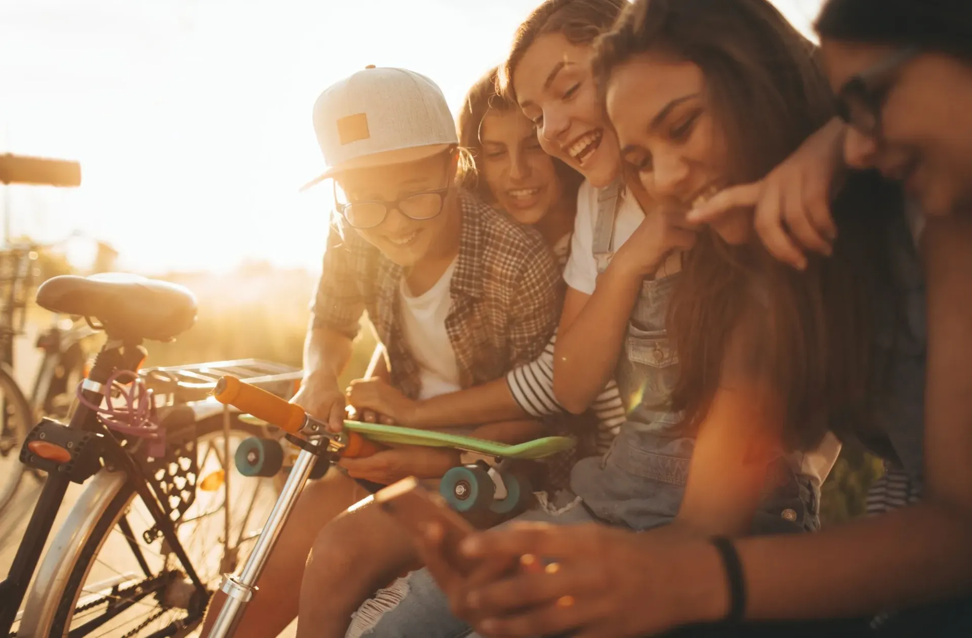 Group of youngpeople laughing, looking at a phone, with skateboards and bikes, outdoors in sunlight.