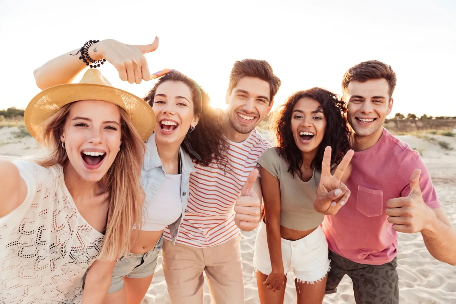 Group of friends smiling and posing outdoors on a sunny beach, giving thumbs up and peace signs.