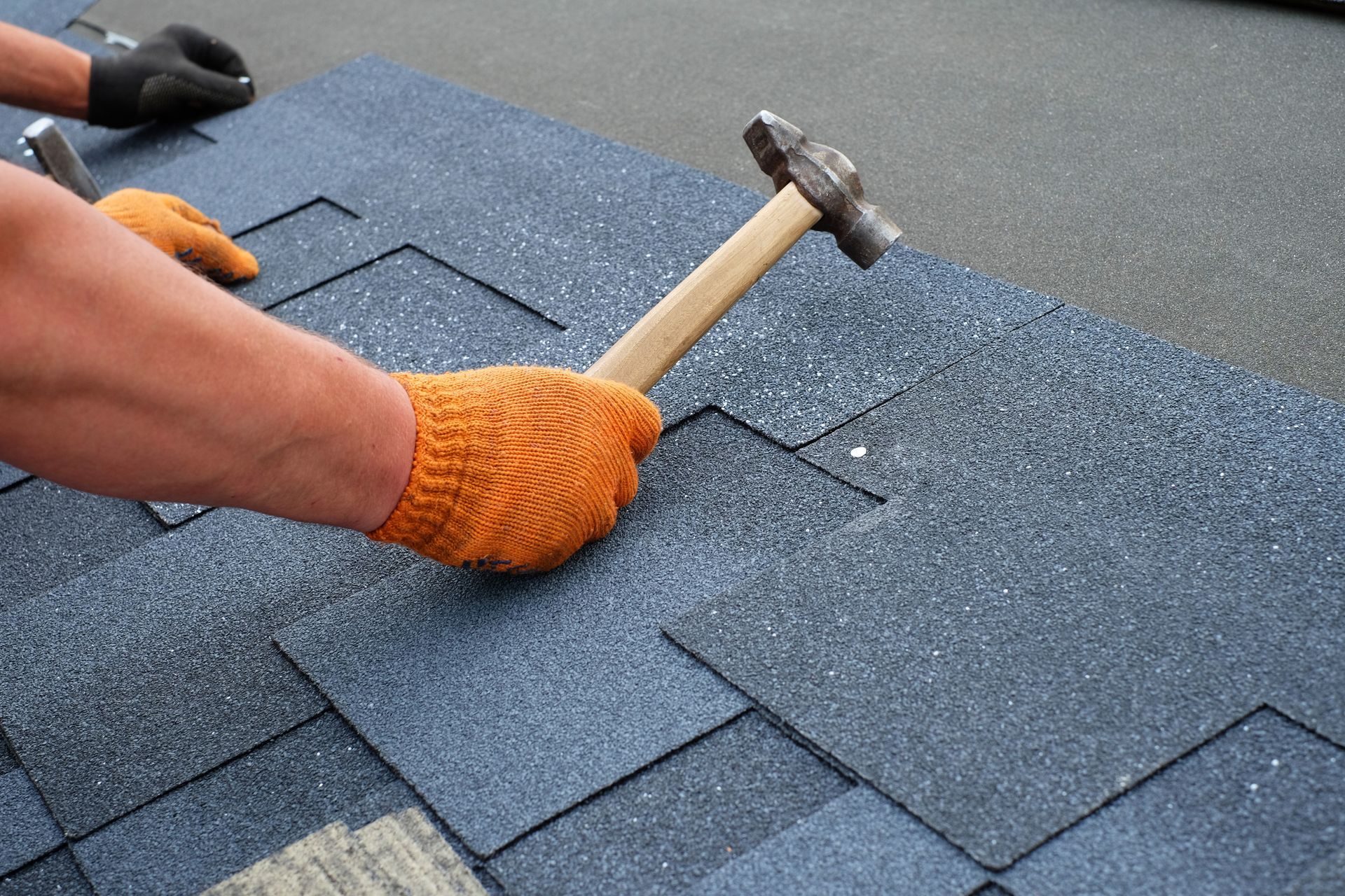 Two workers' hands installing bitumen roof shingles using a hammer.