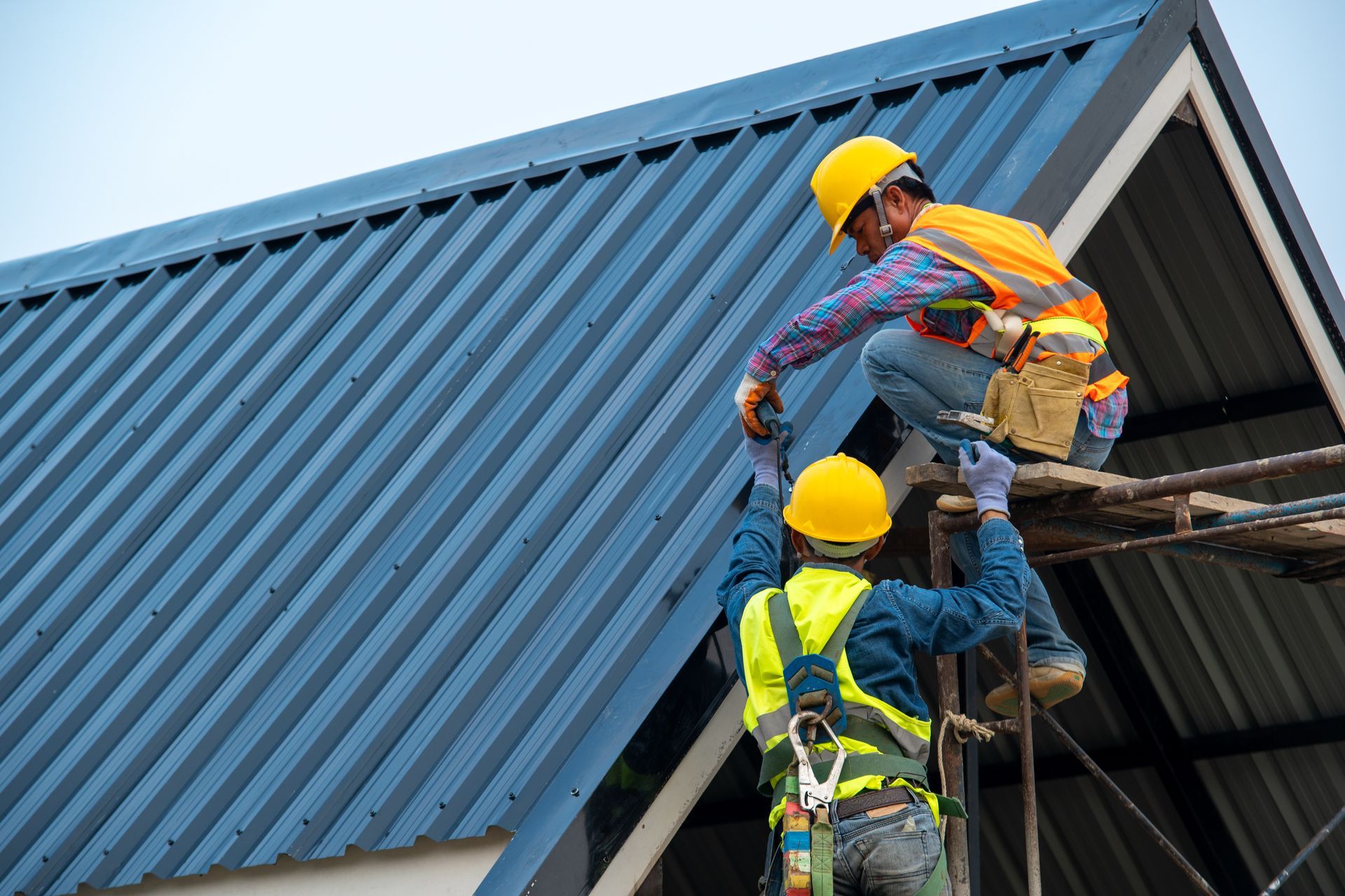 A roofer using an air nail gun while another assists with installing a metal roof.