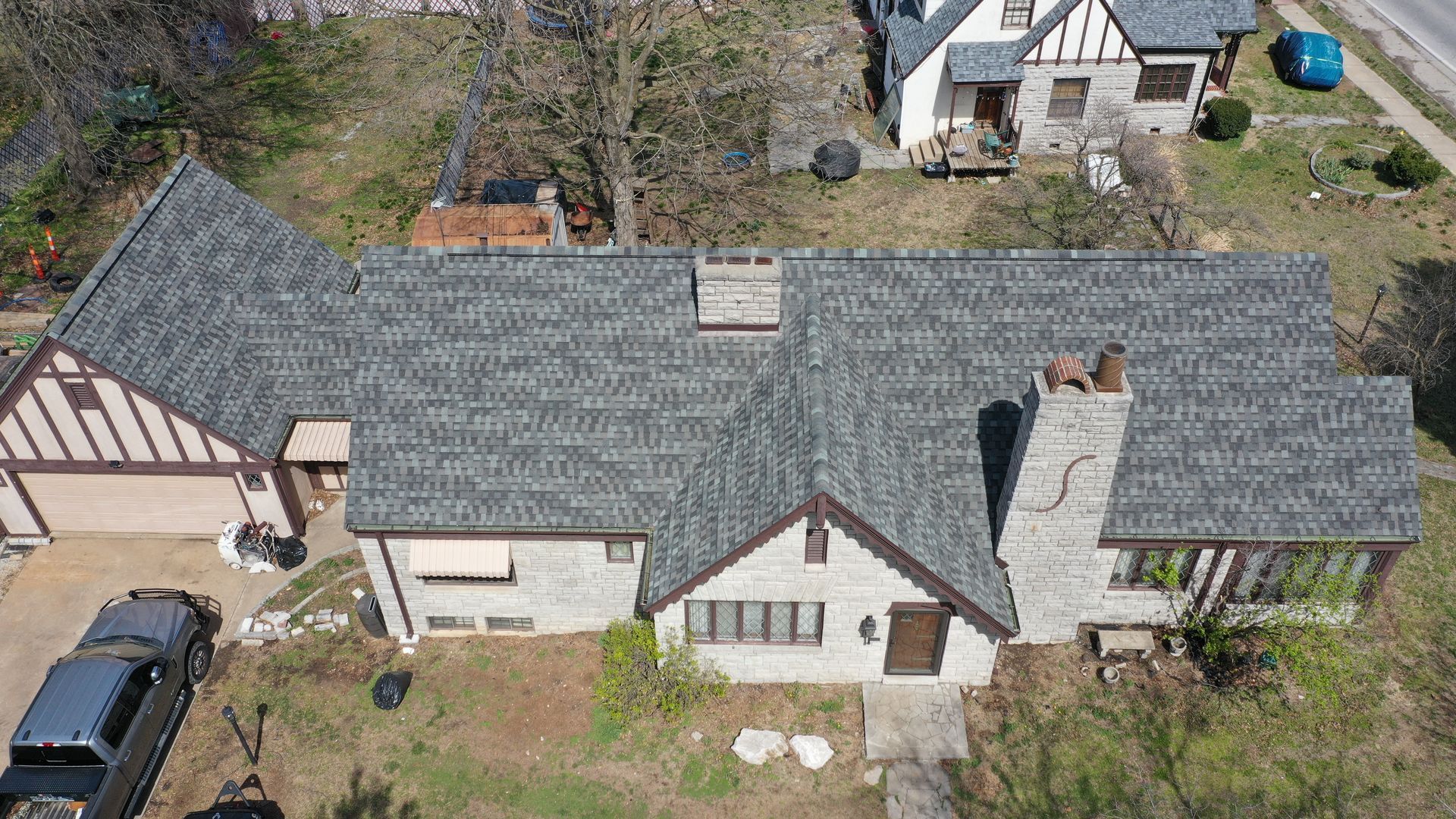 Overhead view of a stone Tudor-style house with a gray roof, chimney, and driveway, alongside a black truck.