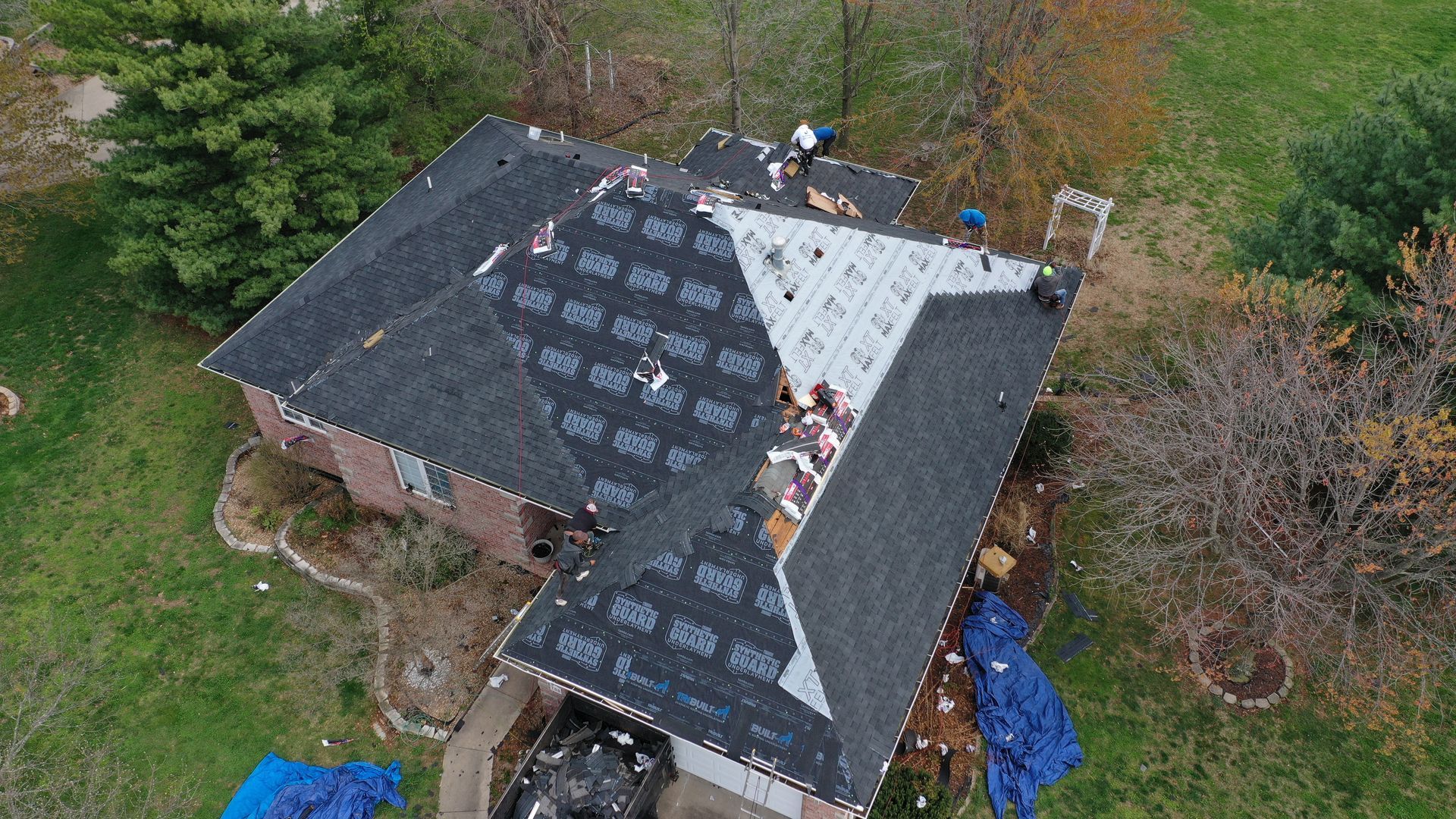 Overhead view of a house with roof damage, being repaired. Dark shingles, blue tarps, and workers are visible.