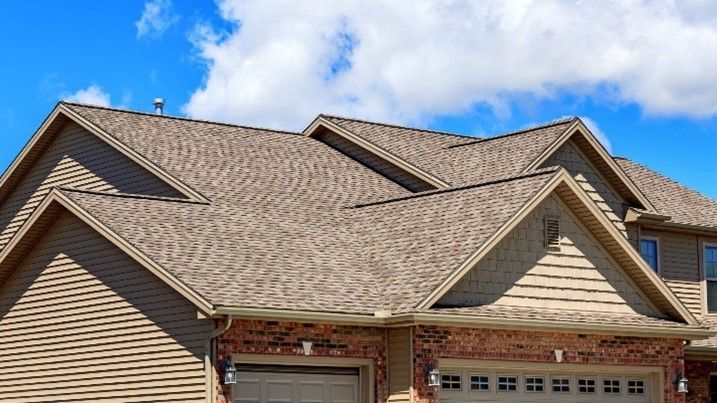 A close-up of a house with beige siding, brick accents, and multiple gable roofs under a blue sky.