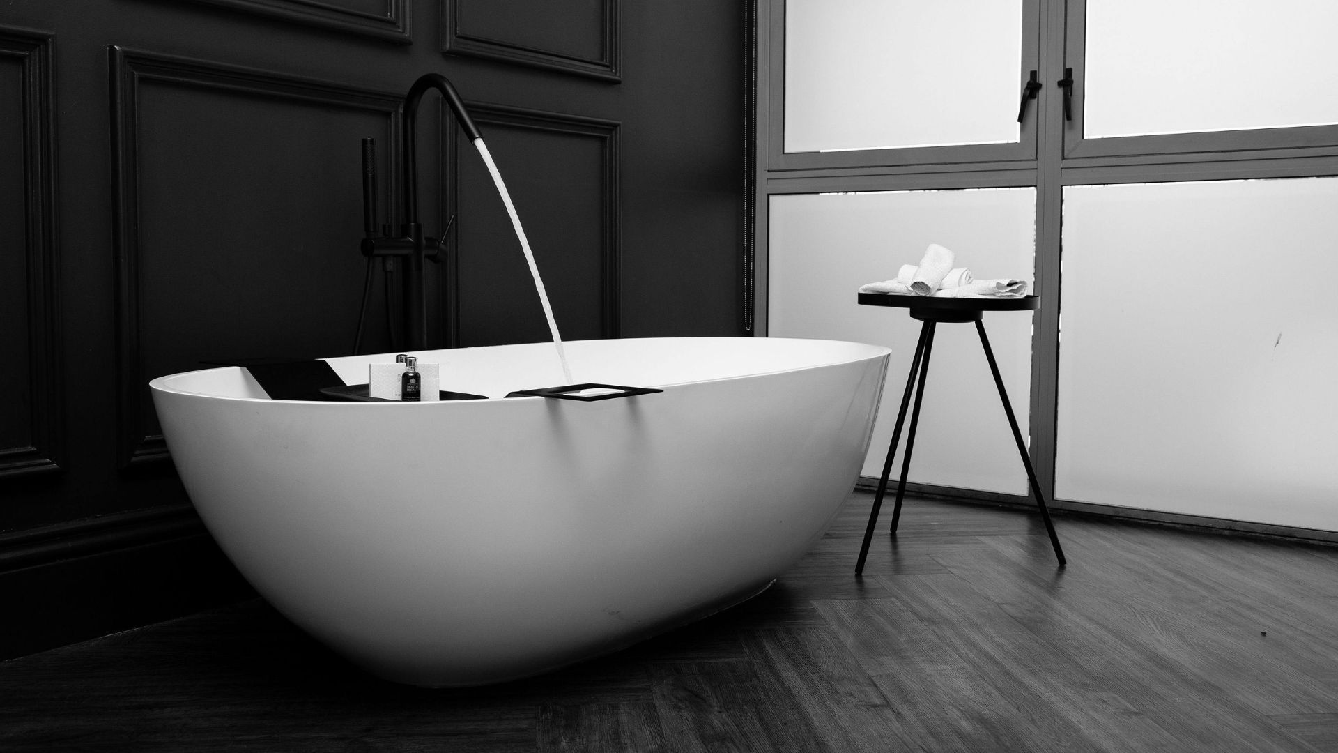 A white, oval freestanding bathtub with a black faucet filling it with water, set against a dark wall and wooden floor.