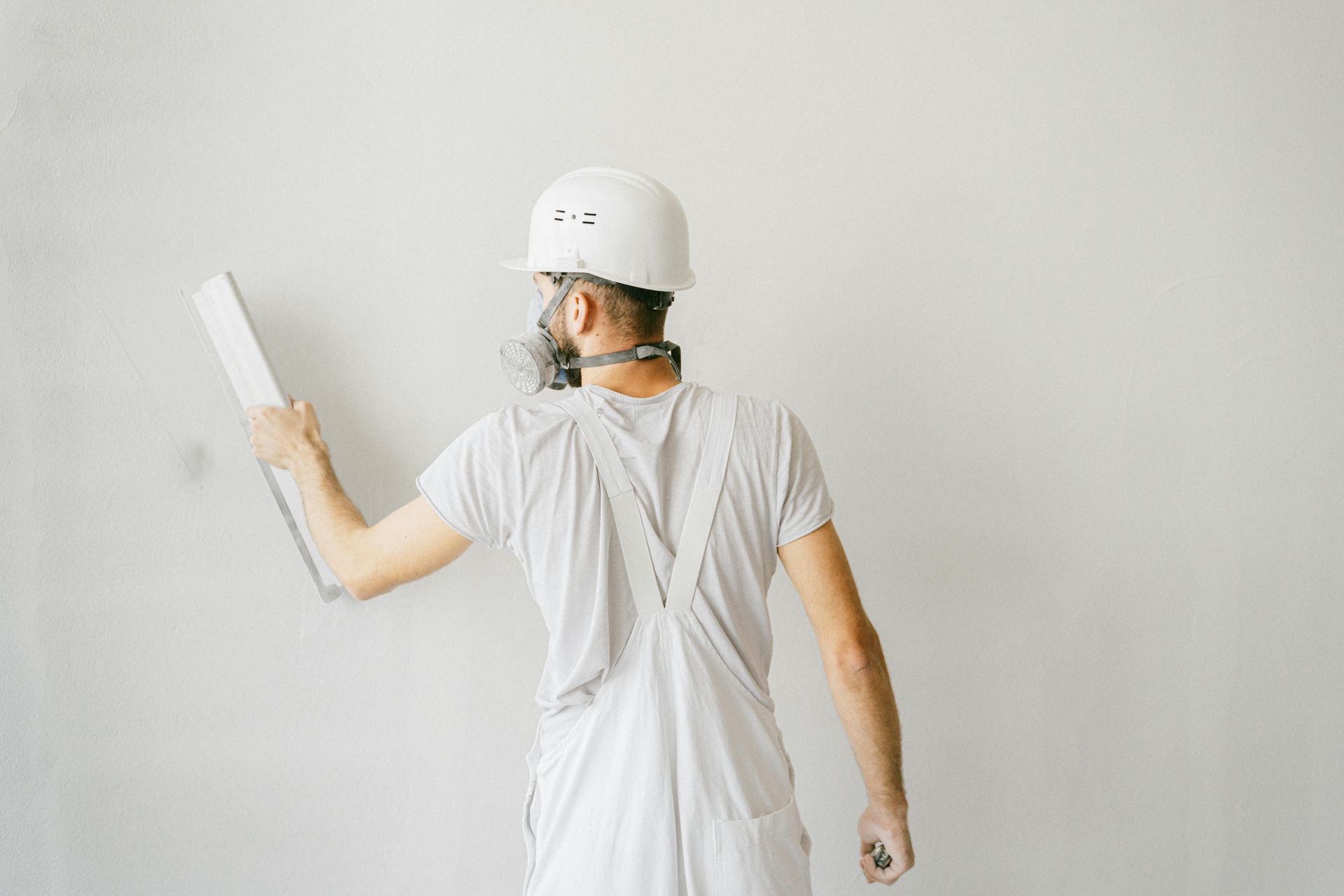 Person in work overalls, hard hat, and respirator uses a trowel to apply compound to a wall.