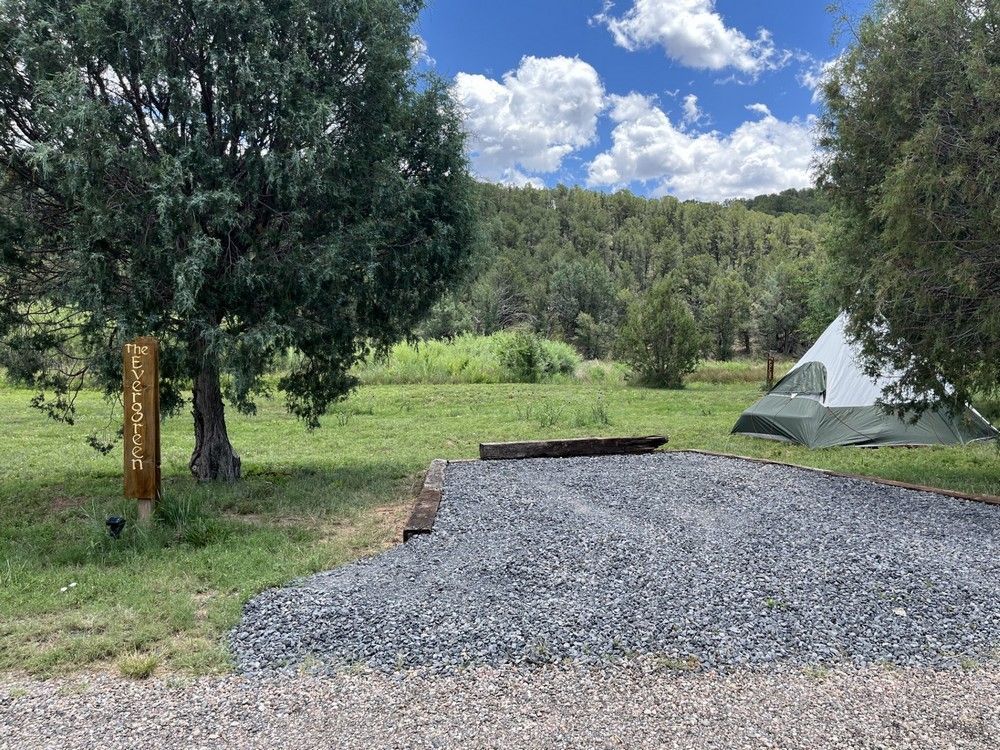 Gravel campsite with tent, sign, and trees against a forested hillside under a blue, cloudy sky.