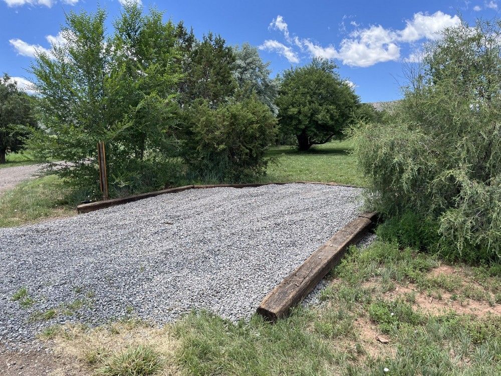 Gravel parking spot bordered by wood beams, surrounded by grass and trees under a blue sky.