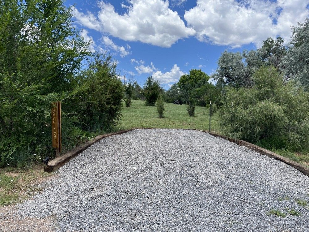 Gravel driveway leading into a grassy field with trees under a blue sky with clouds.
