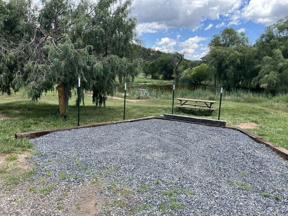 Gravel campsite with picnic table, surrounded by trees and a pond, under a cloudy sky.