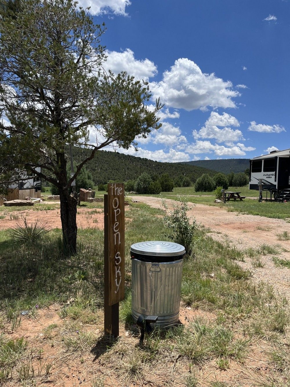 Metal trash can and wooden sign in a campsite with trees and mountains under a blue sky with clouds.