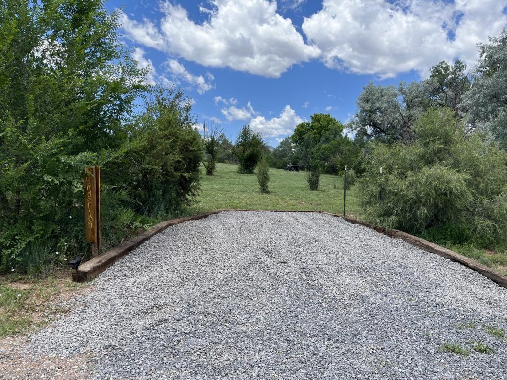 Gravel driveway leading to a grassy field bordered by trees under a blue sky with clouds.