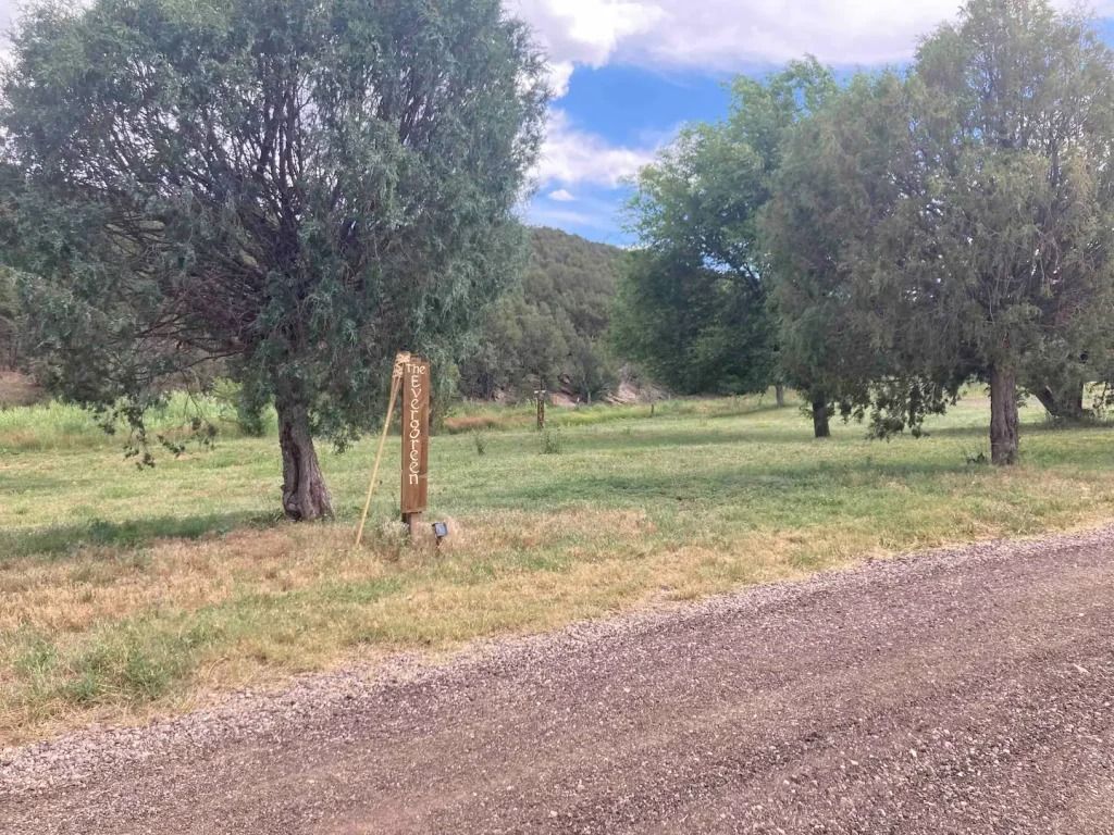 Gravel road leads to a grassy area with trees and a wooden sign under a cloudy sky.