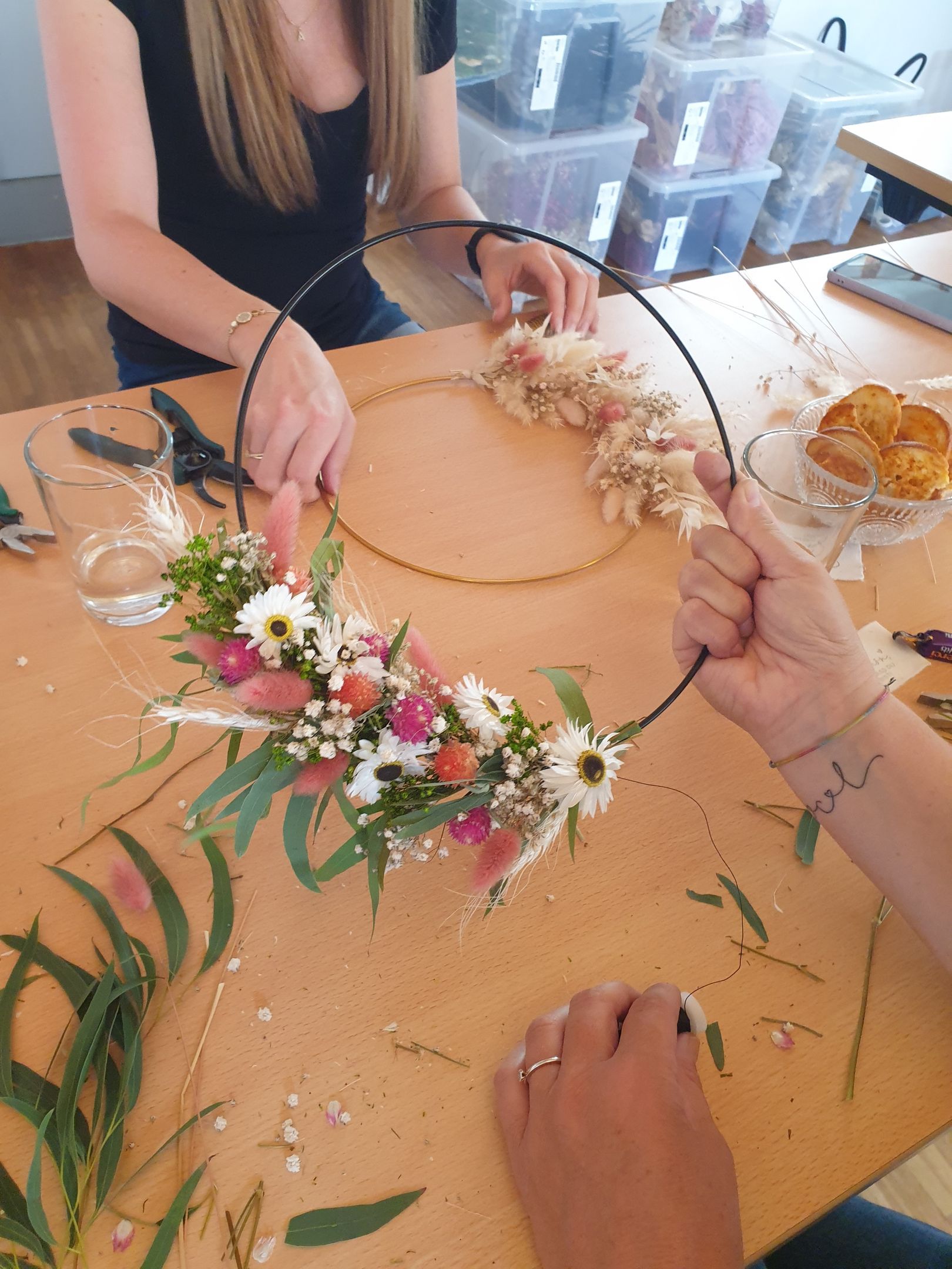 Two women are sitting at a table making a flower crown.