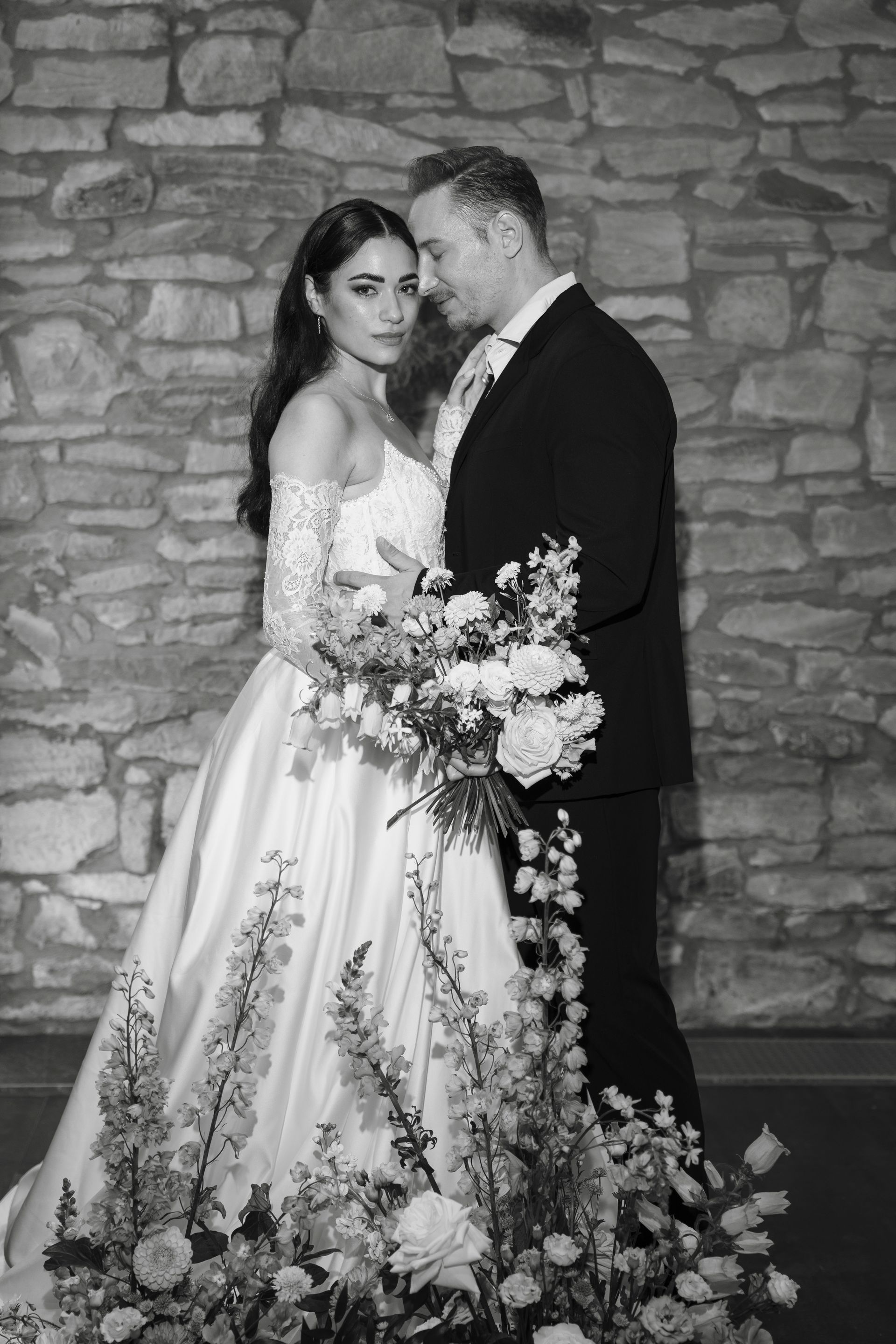 A bride and groom are posing for a black and white photo in front of a stone wall.