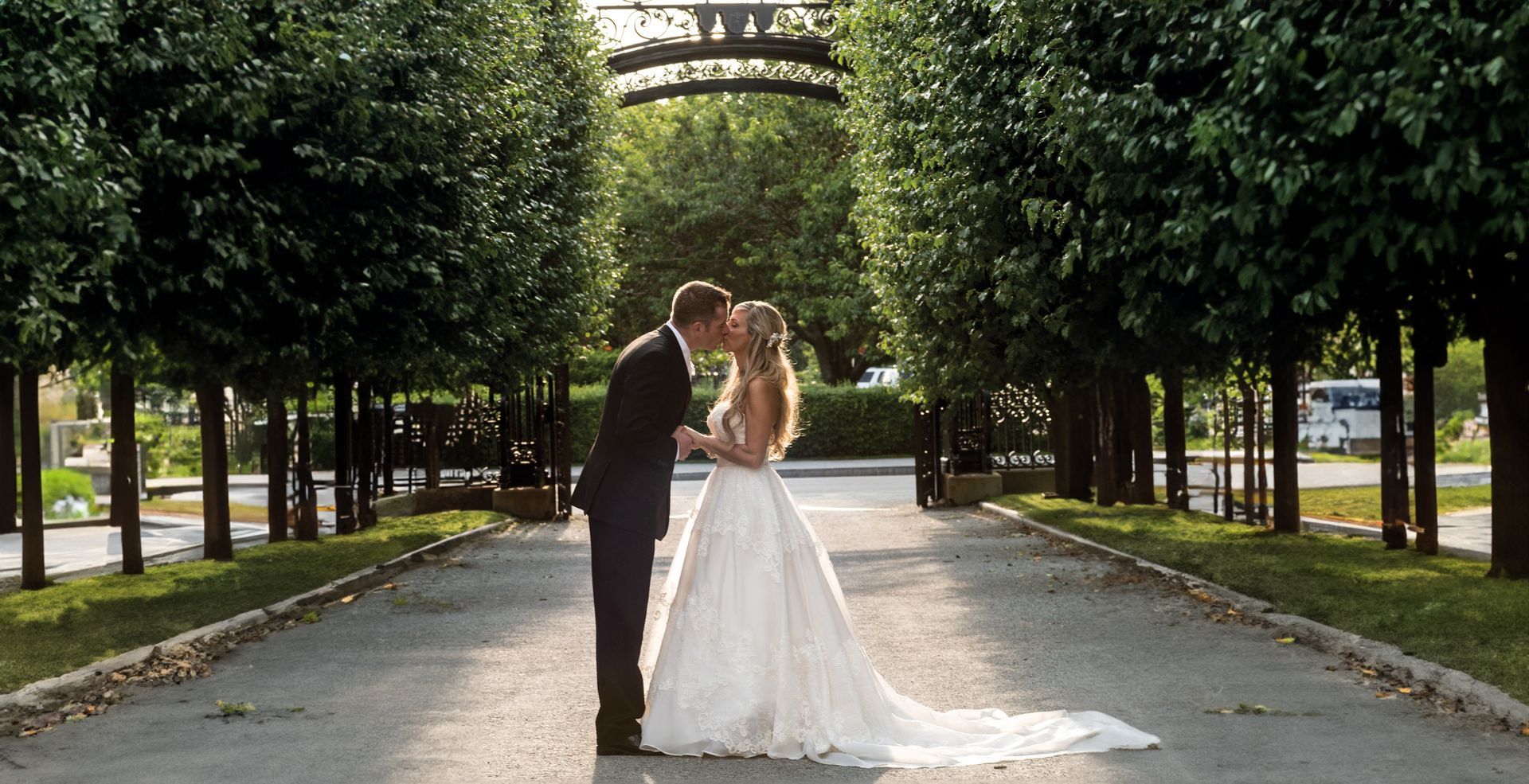 A bride and groom are kissing on a path lined with trees.