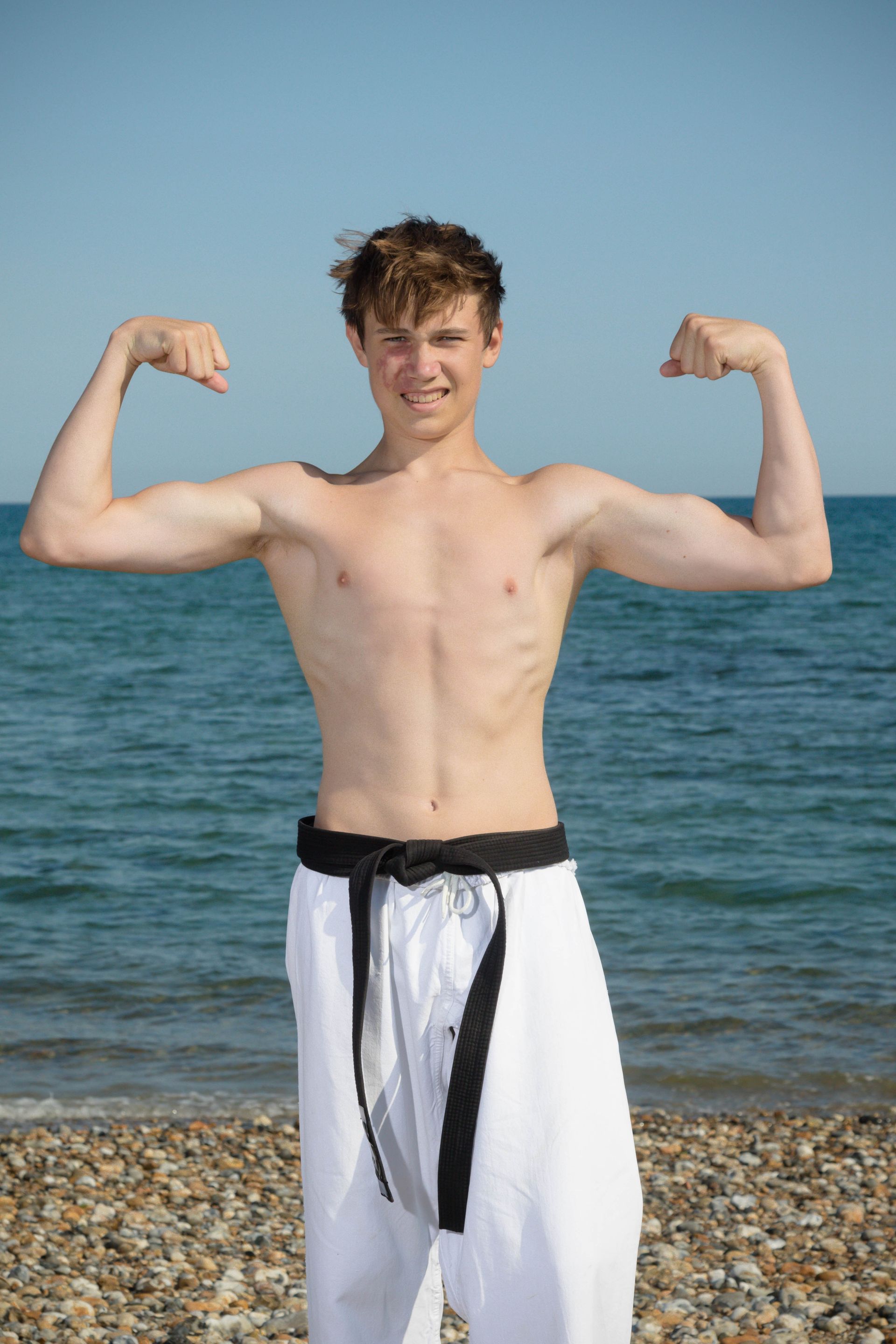 A shirtless young man is flexing his muscles on the beach.