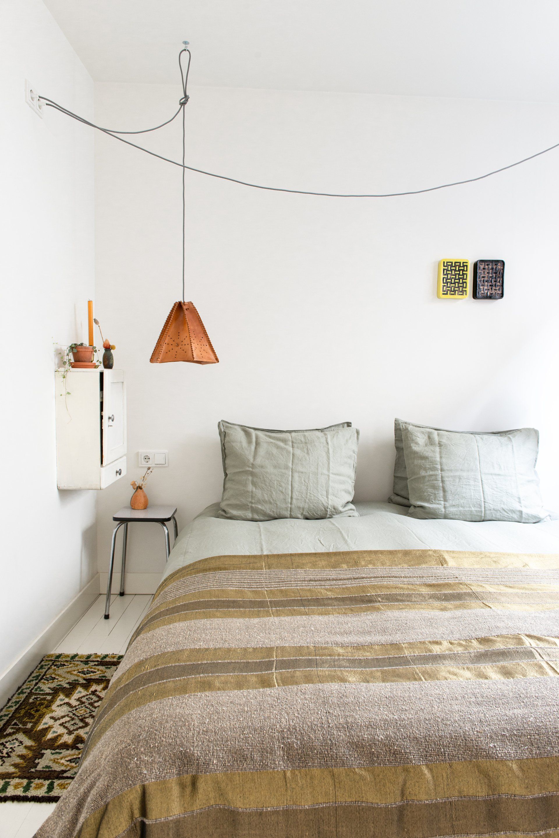 Slaapkamer met een witte muur, bed met bruine deken en kussens, koperen hanglamp en een klein bijzettafeltje.