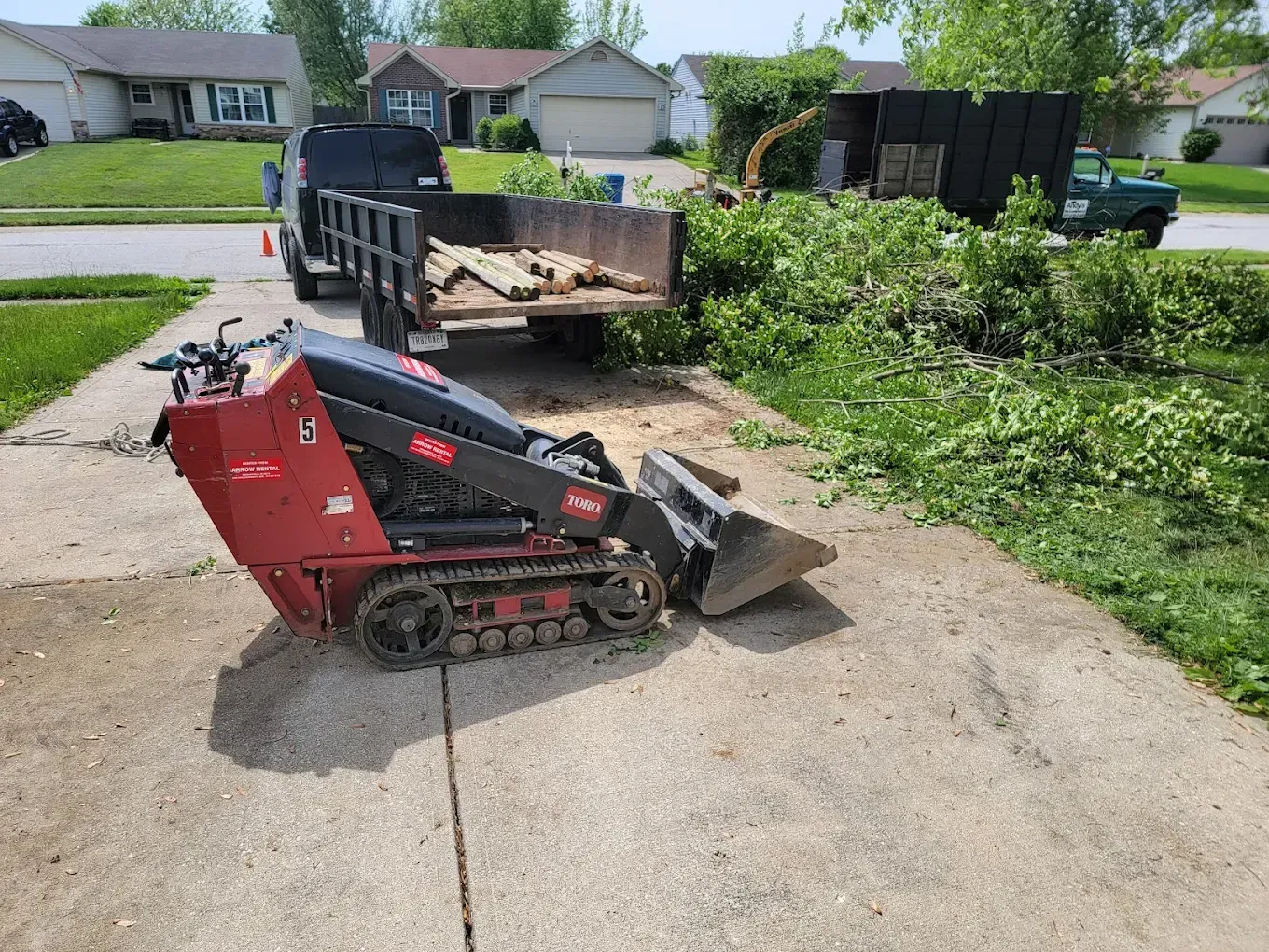 Red skid-steer loader beside a dump trailer on a suburban driveway with brush piles nearby