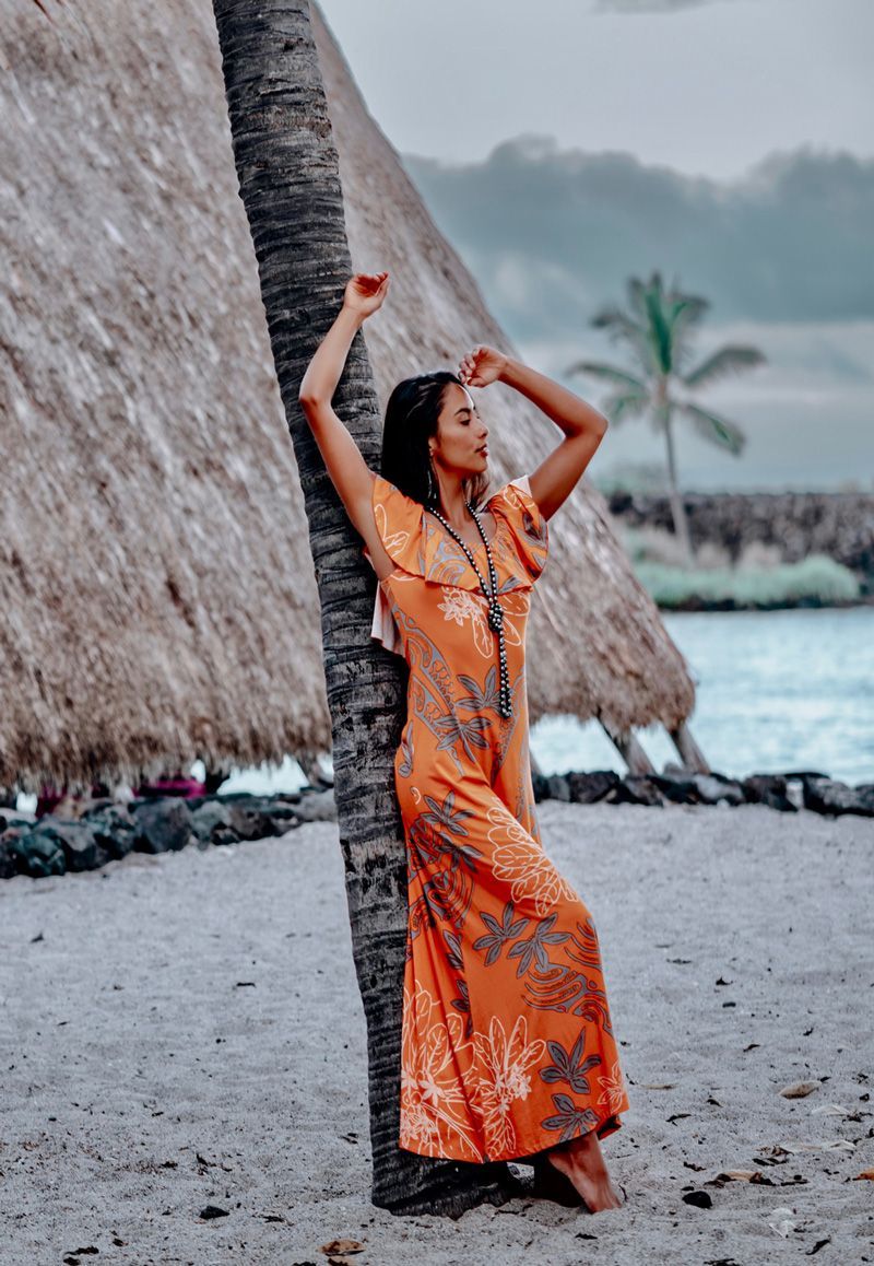 A woman in an orange dress is leaning against a palm tree on the beach.