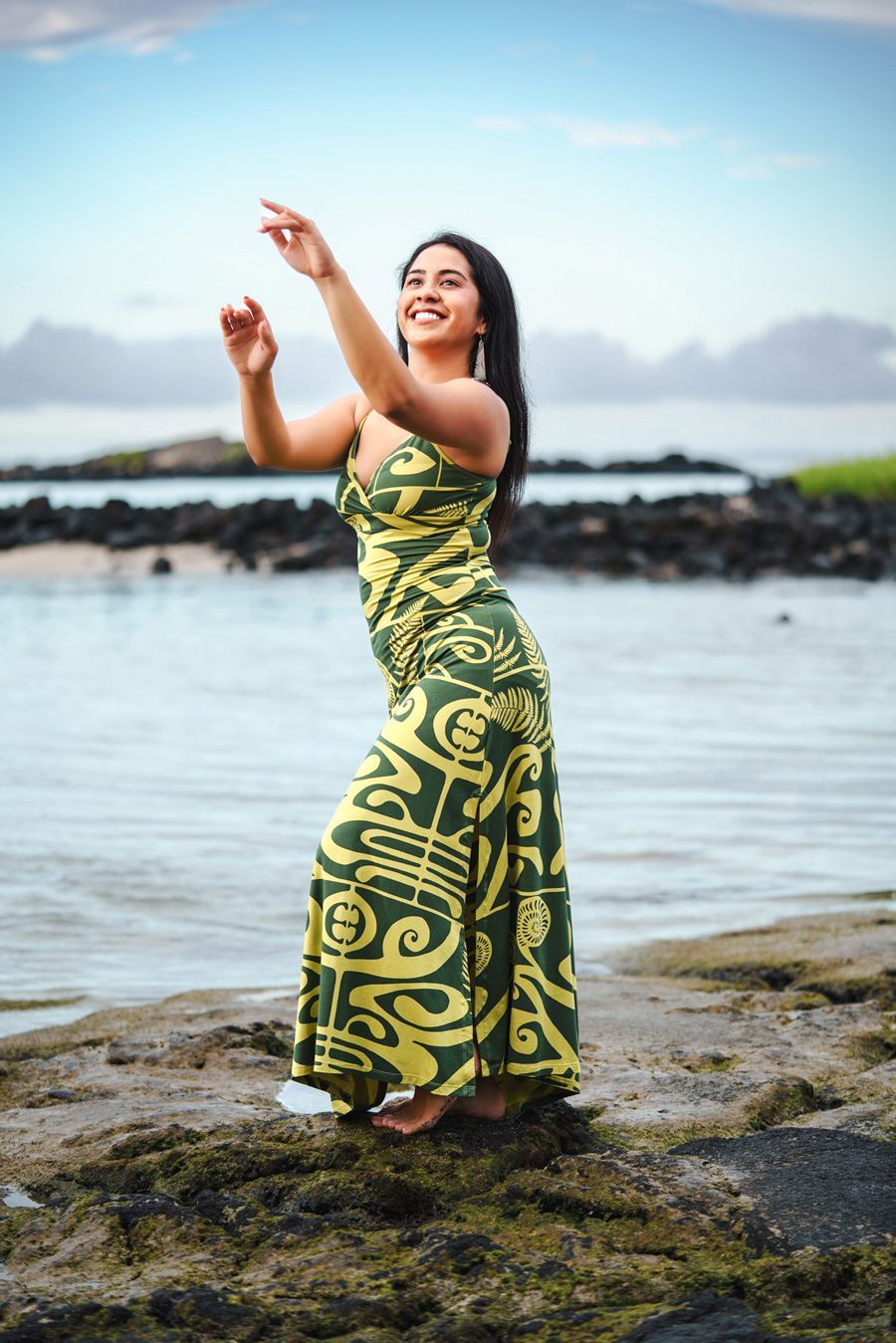 A woman in a green and yellow dress is standing on a rock near the water.