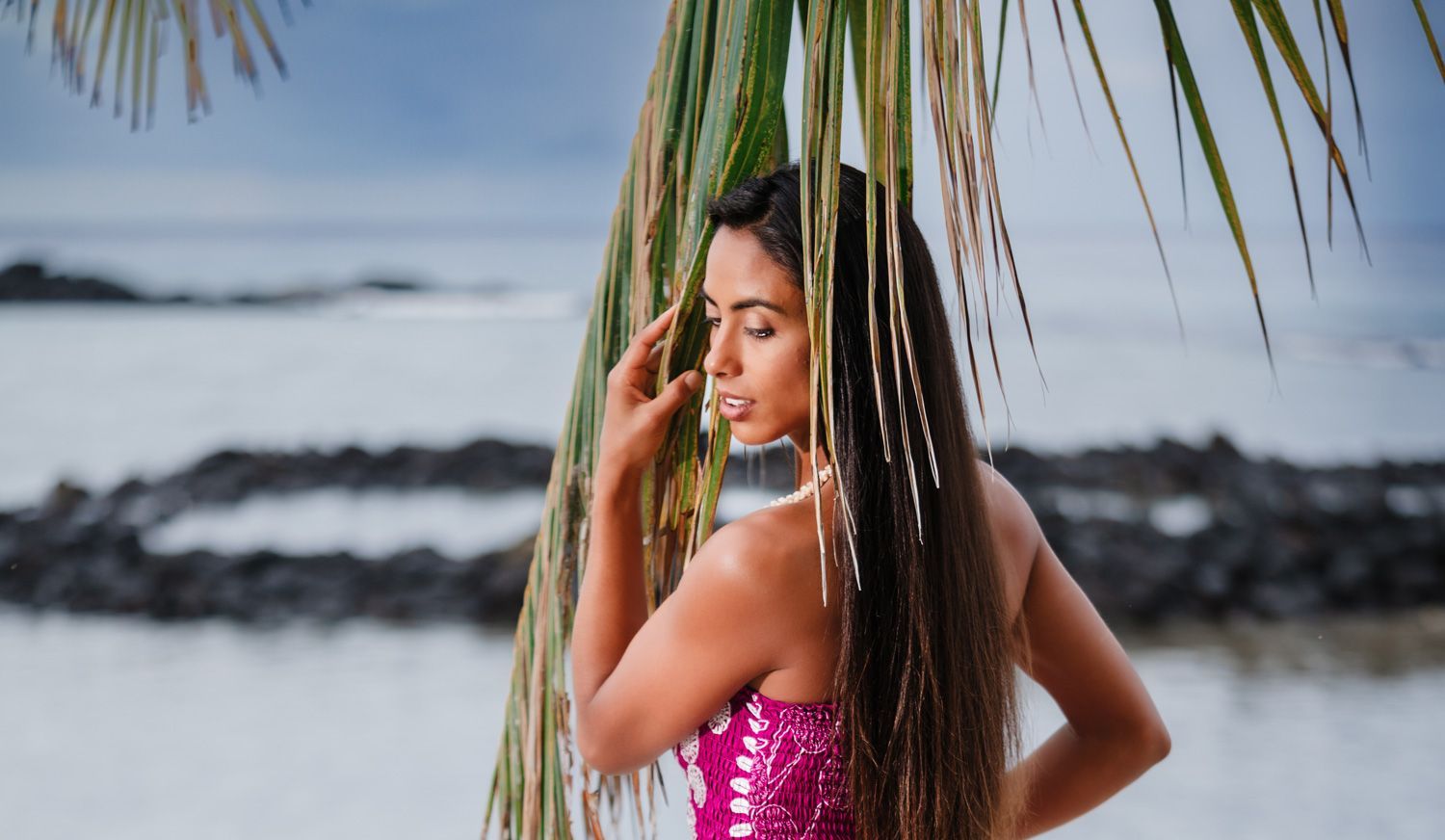 A woman in a pink dress is standing next to a palm tree on the beach.