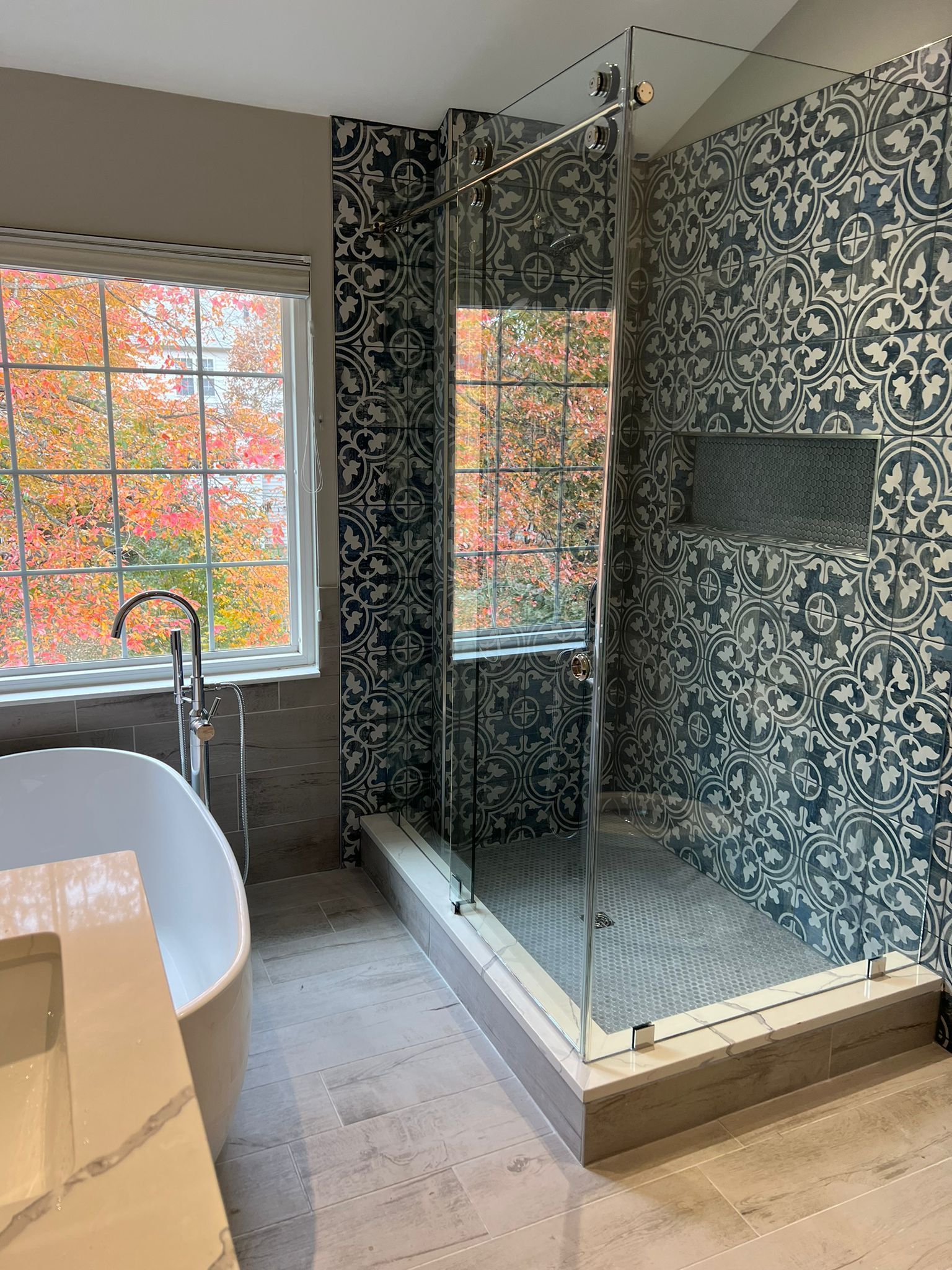 Bathroom with a glass shower, patterned blue and white tile, and a window with fall foliage.