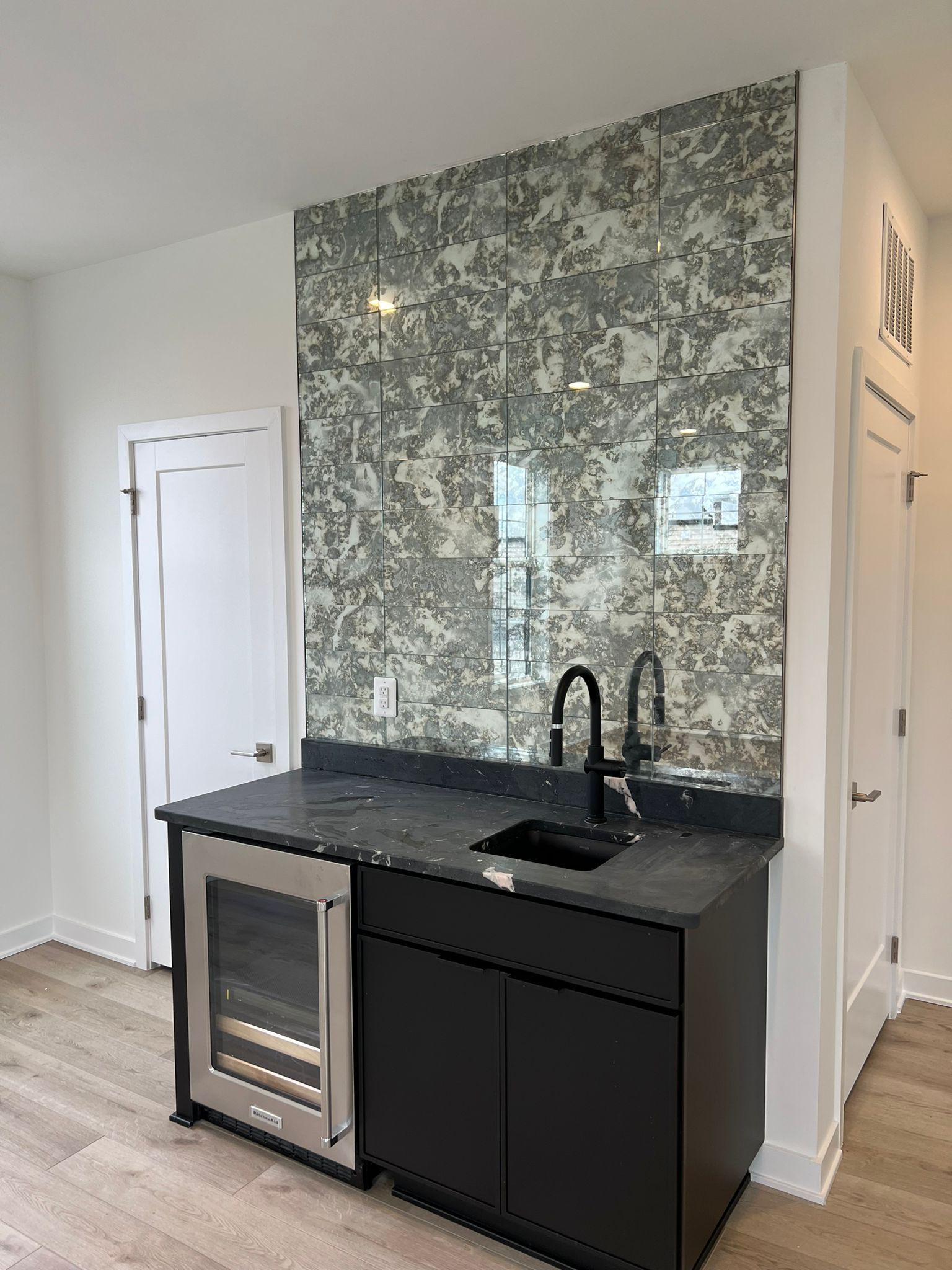 Small black bar with wine fridge, sink, and textured mirror backsplash.