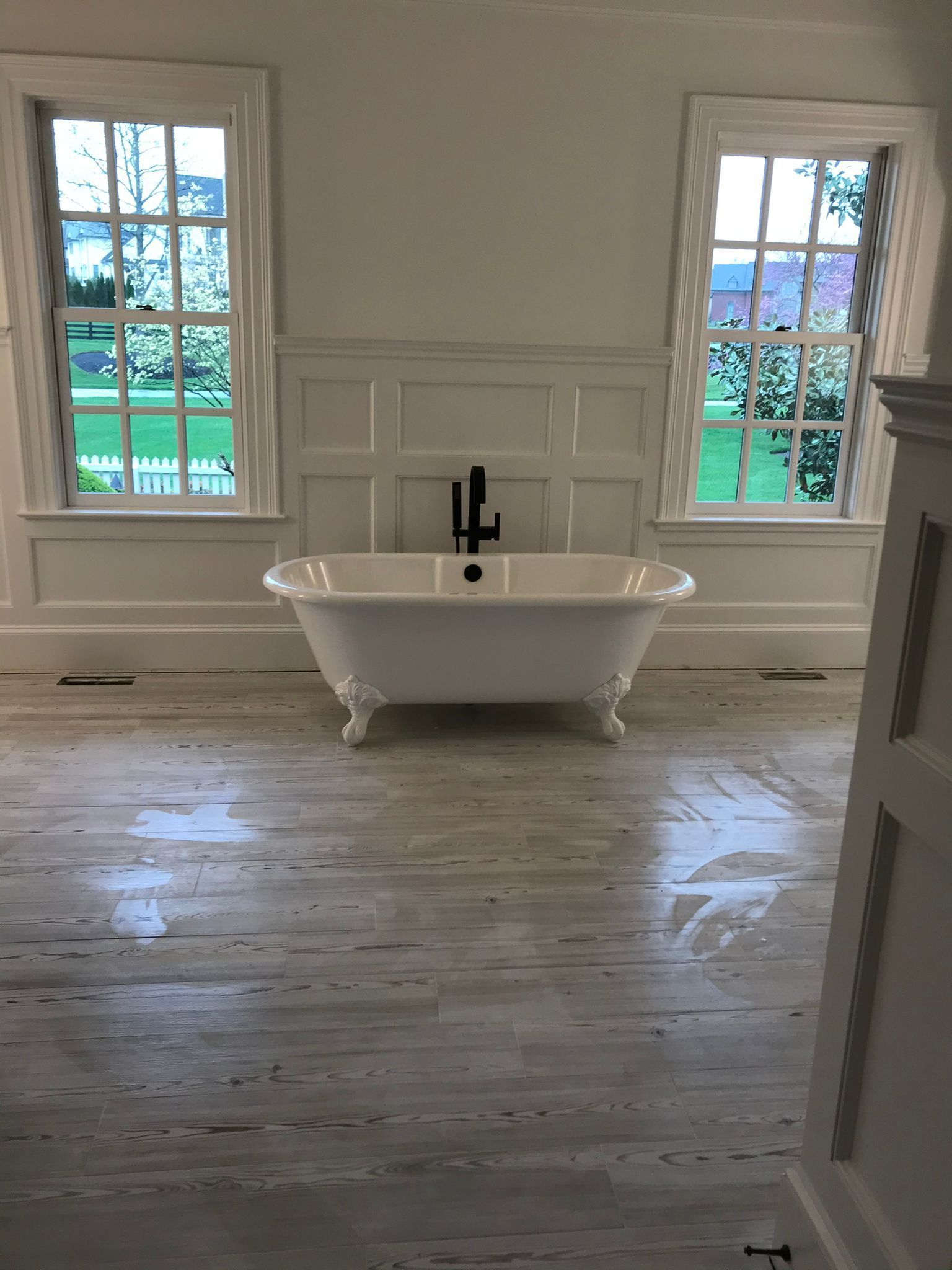 Clawfoot bathtub centered between two windows in a white-walled room with wood floors.