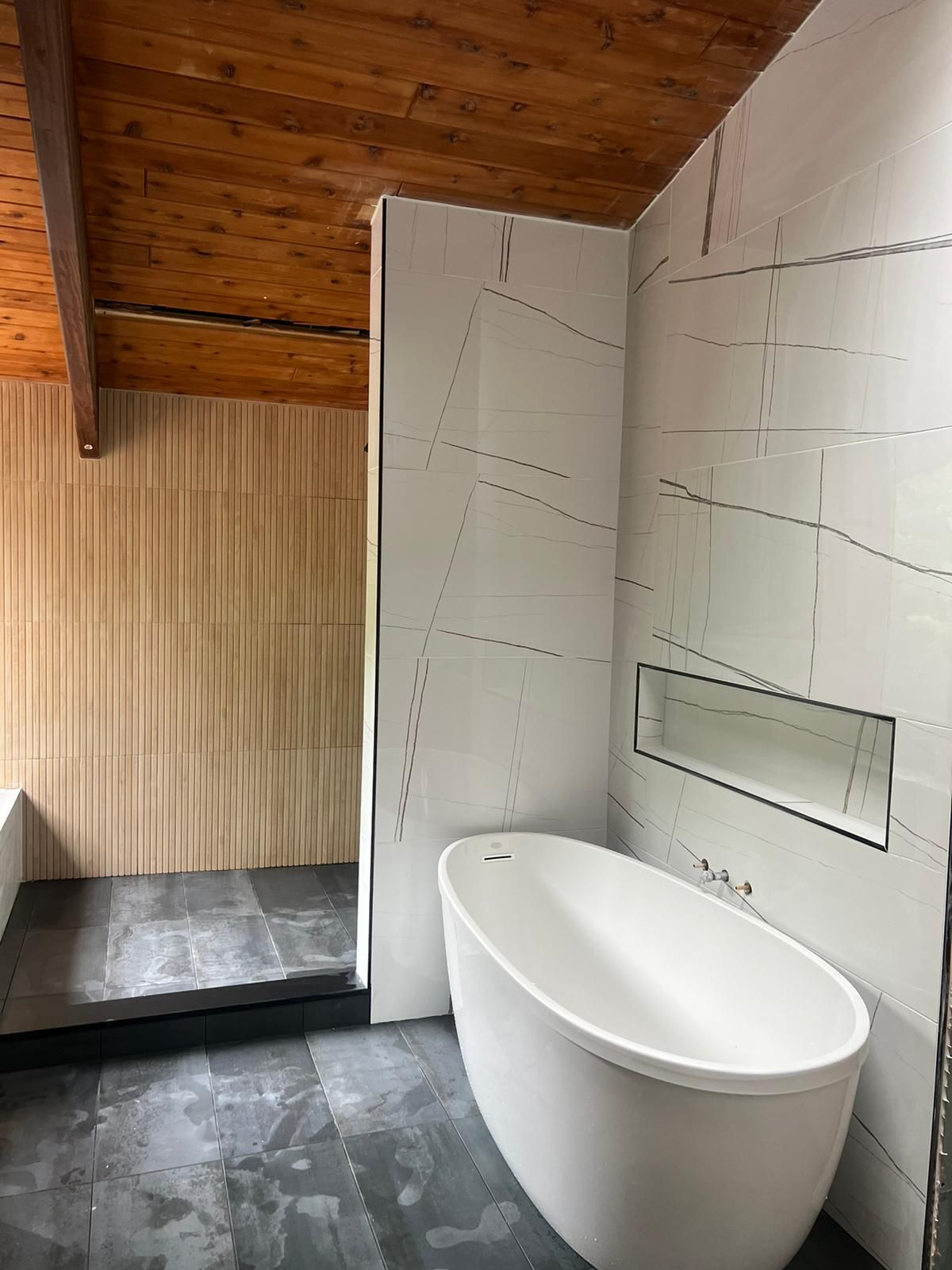 Bathroom with white soaking tub, white and grey tiled wall, and wood ceiling.