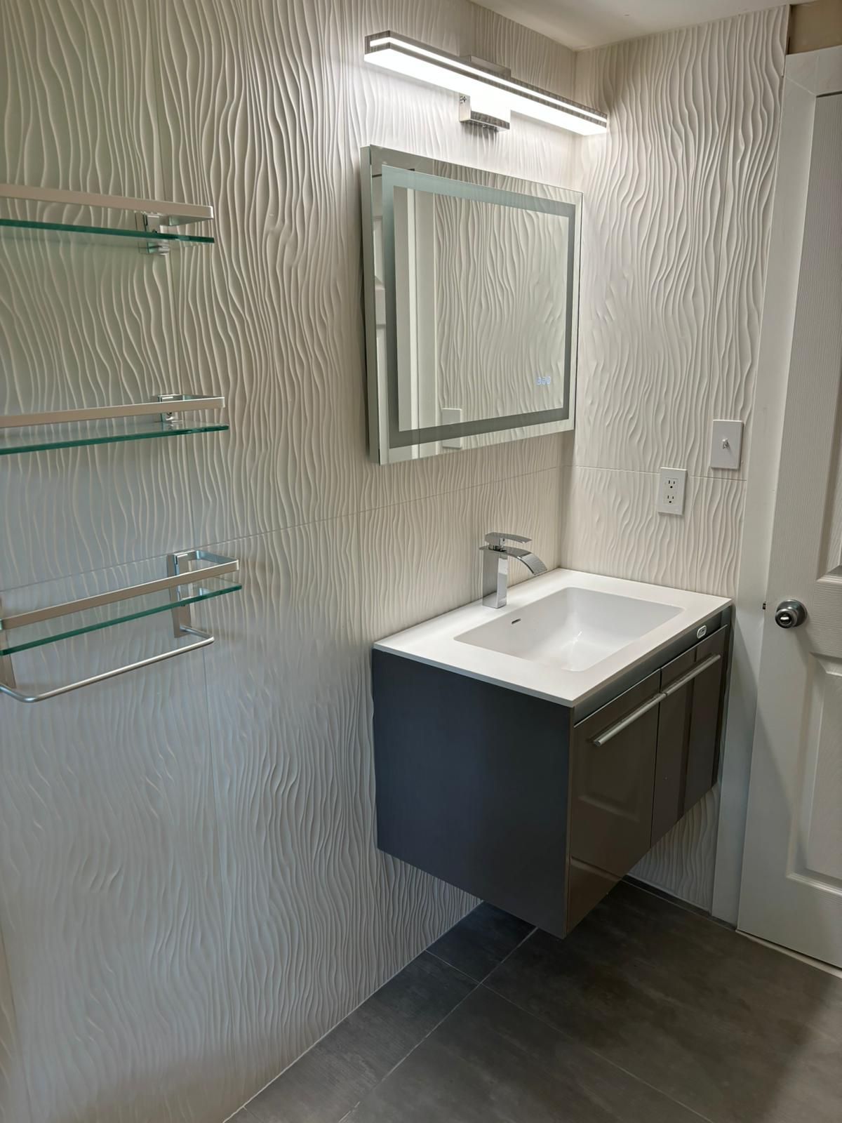 Modern bathroom with a floating vanity, mirror, and glass shelves against textured wallpaper.