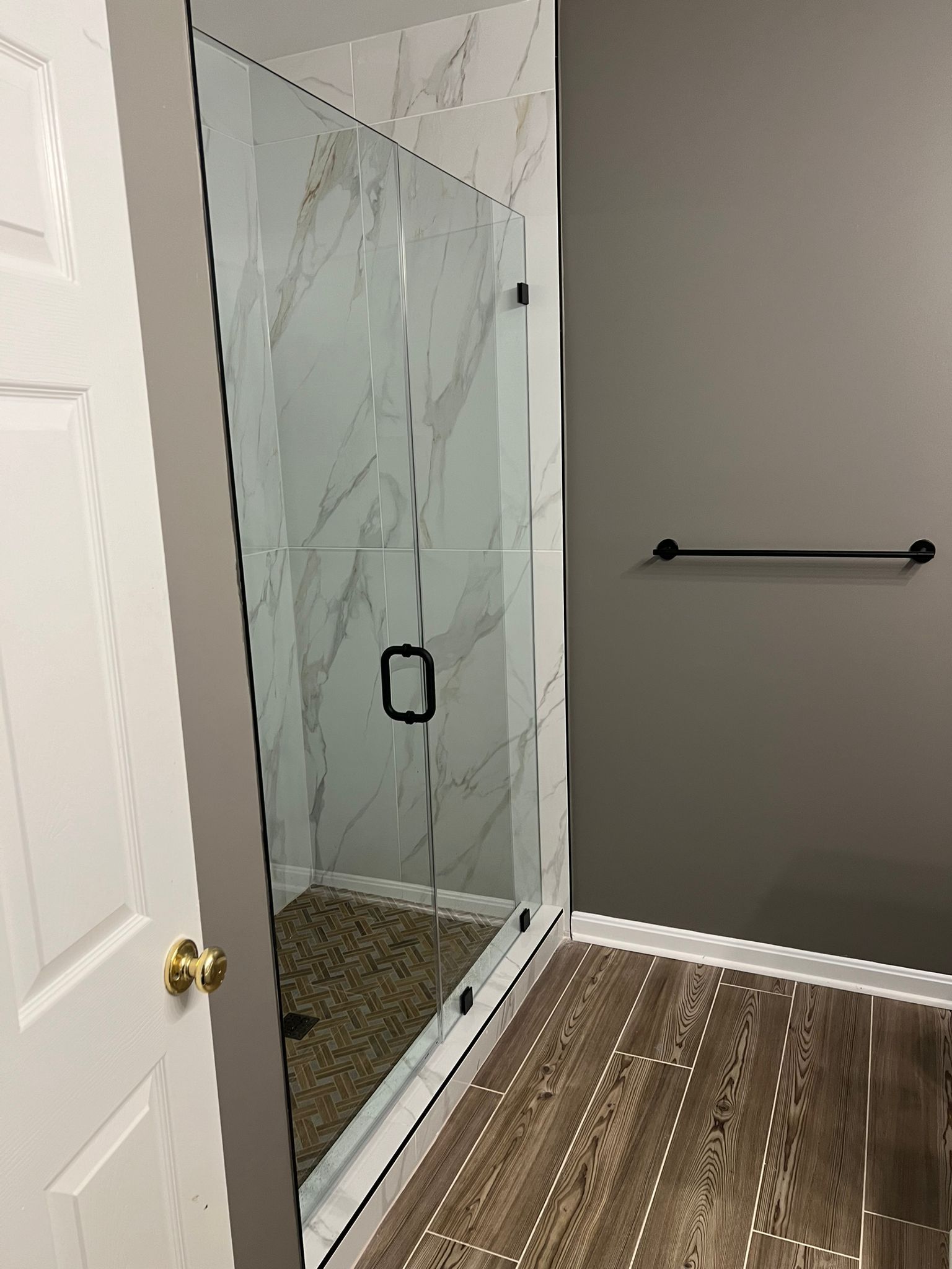 Shower with glass door, marble tile walls, and stone floor. Dark towel rack on gray wall, light wood floor.