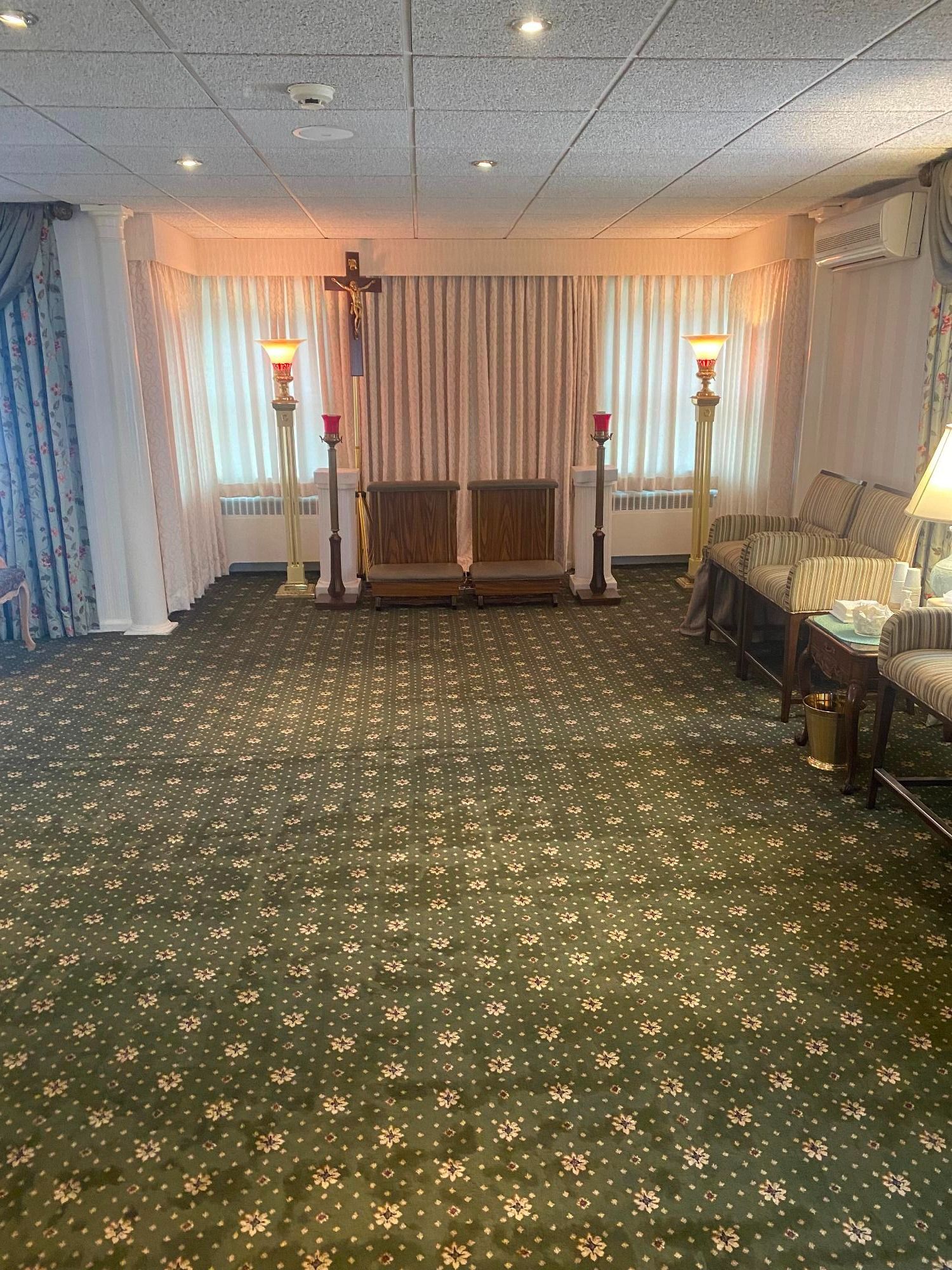 Funeral home interior with chairs, cross, and decorative lighting. Green carpet.