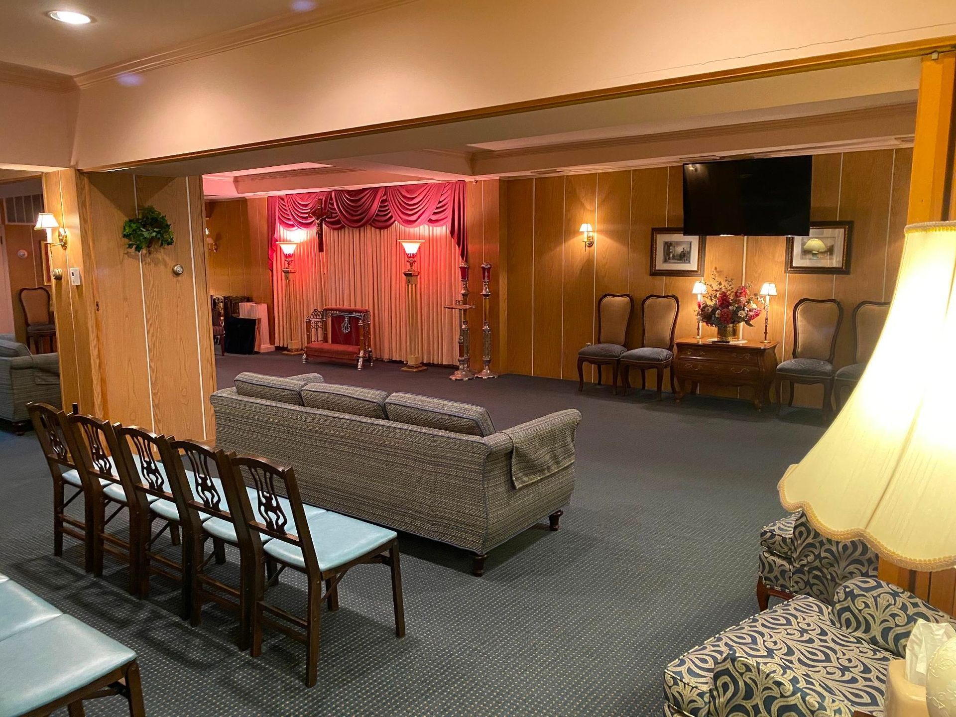 Funeral home interior; chairs, couches, floral arrangements, stage with curtains. Soft lighting, neutral colors.