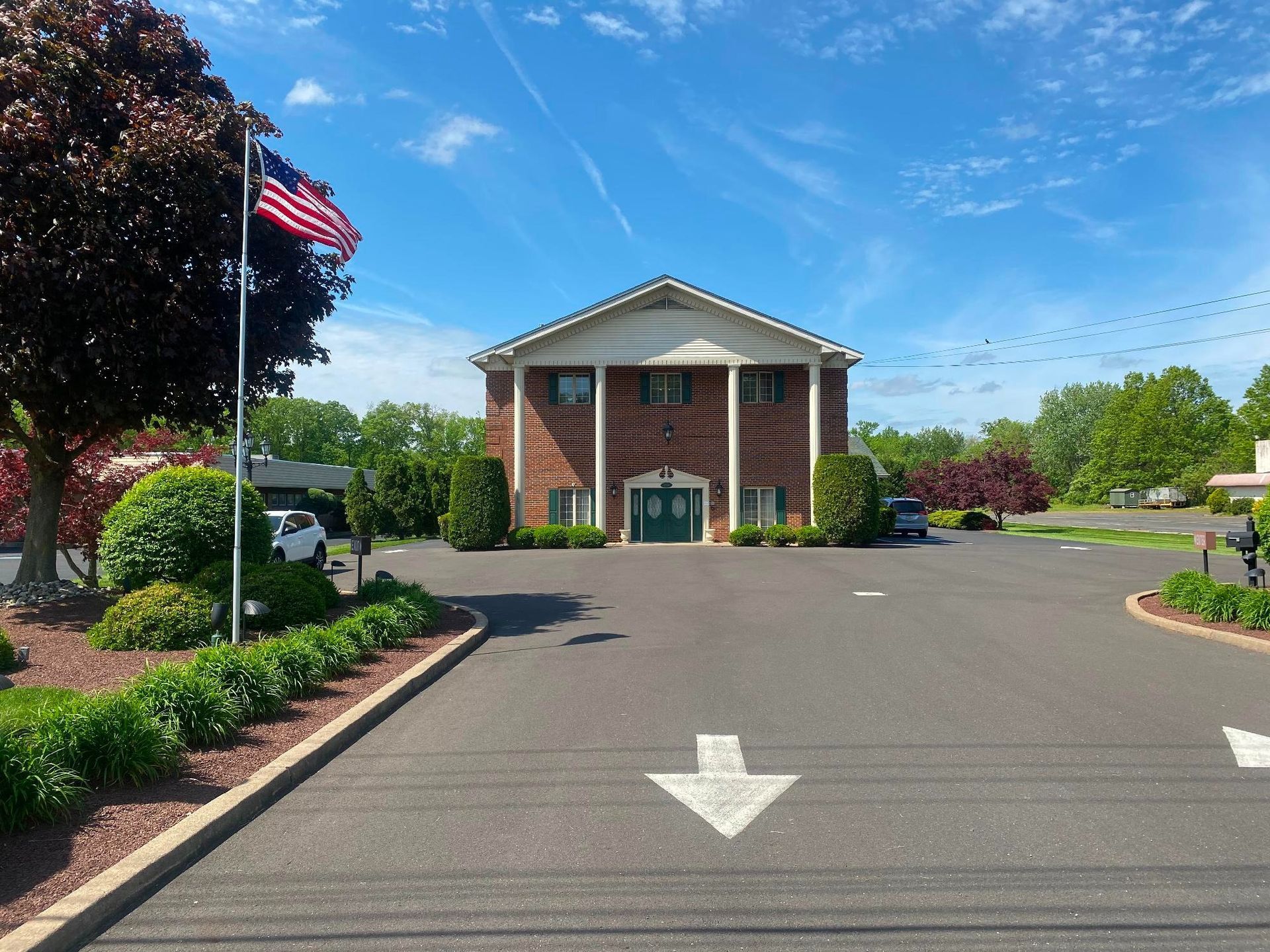 Two-story brick building with white columns and green door; American flag flies in front. Driveway with arrow.