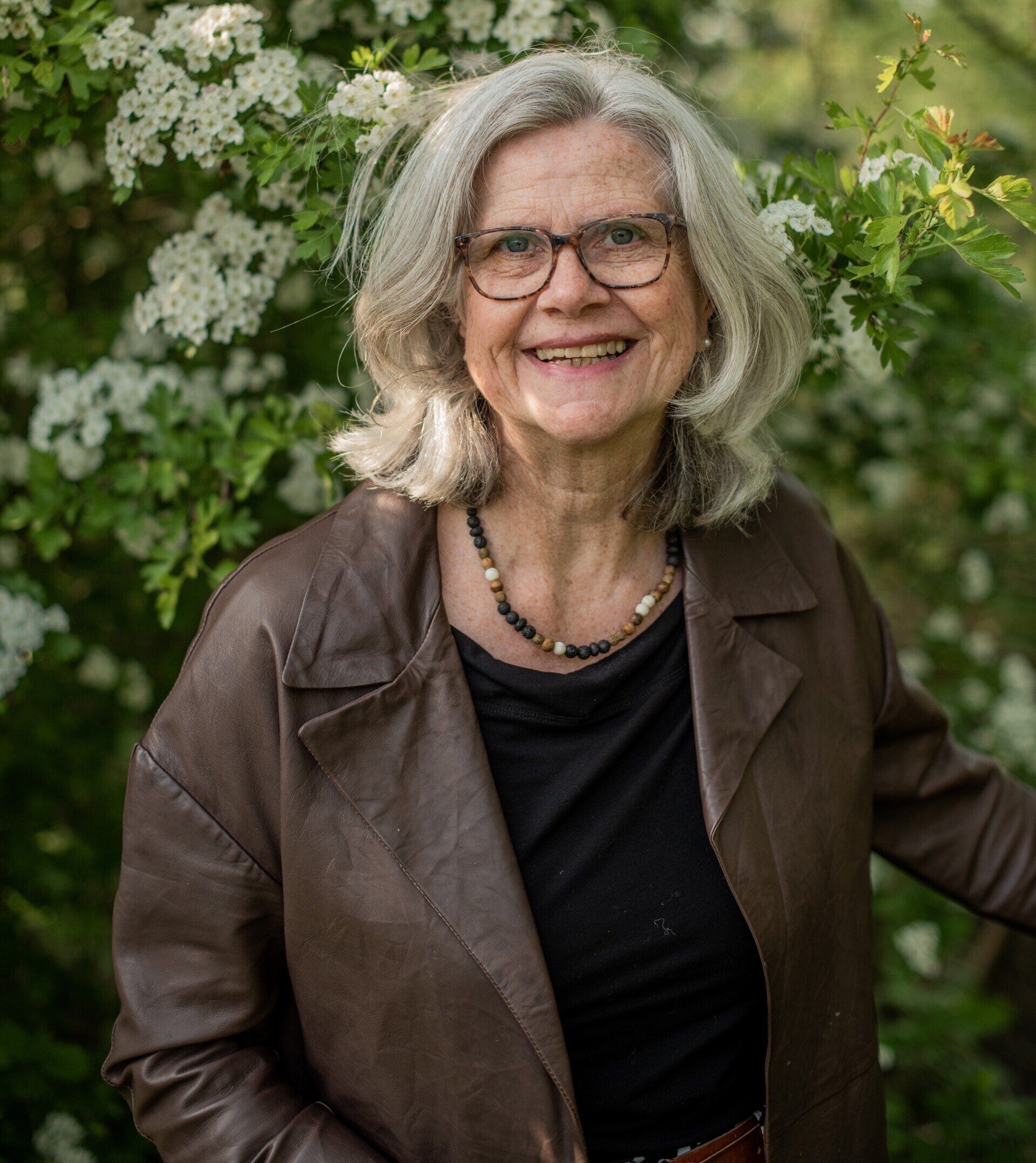 Een vrouw met een bril en een leren jas lacht voor witte bloemen.