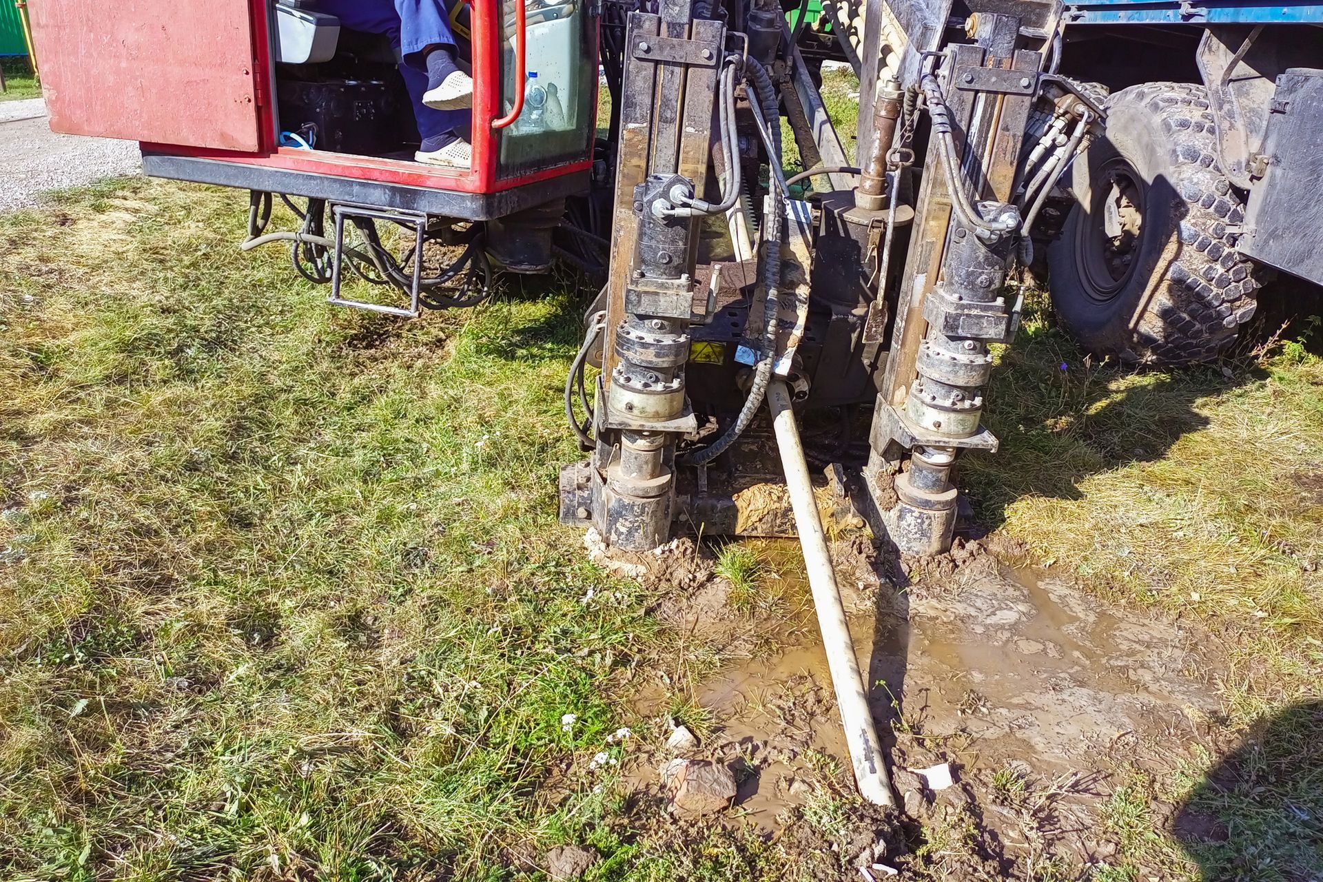 A close-up of a drilling machine on grass with pipes, mud, and the operator partially visible A close-up of a drilling machine on grass with pipes, mud, and the operator partially visible
