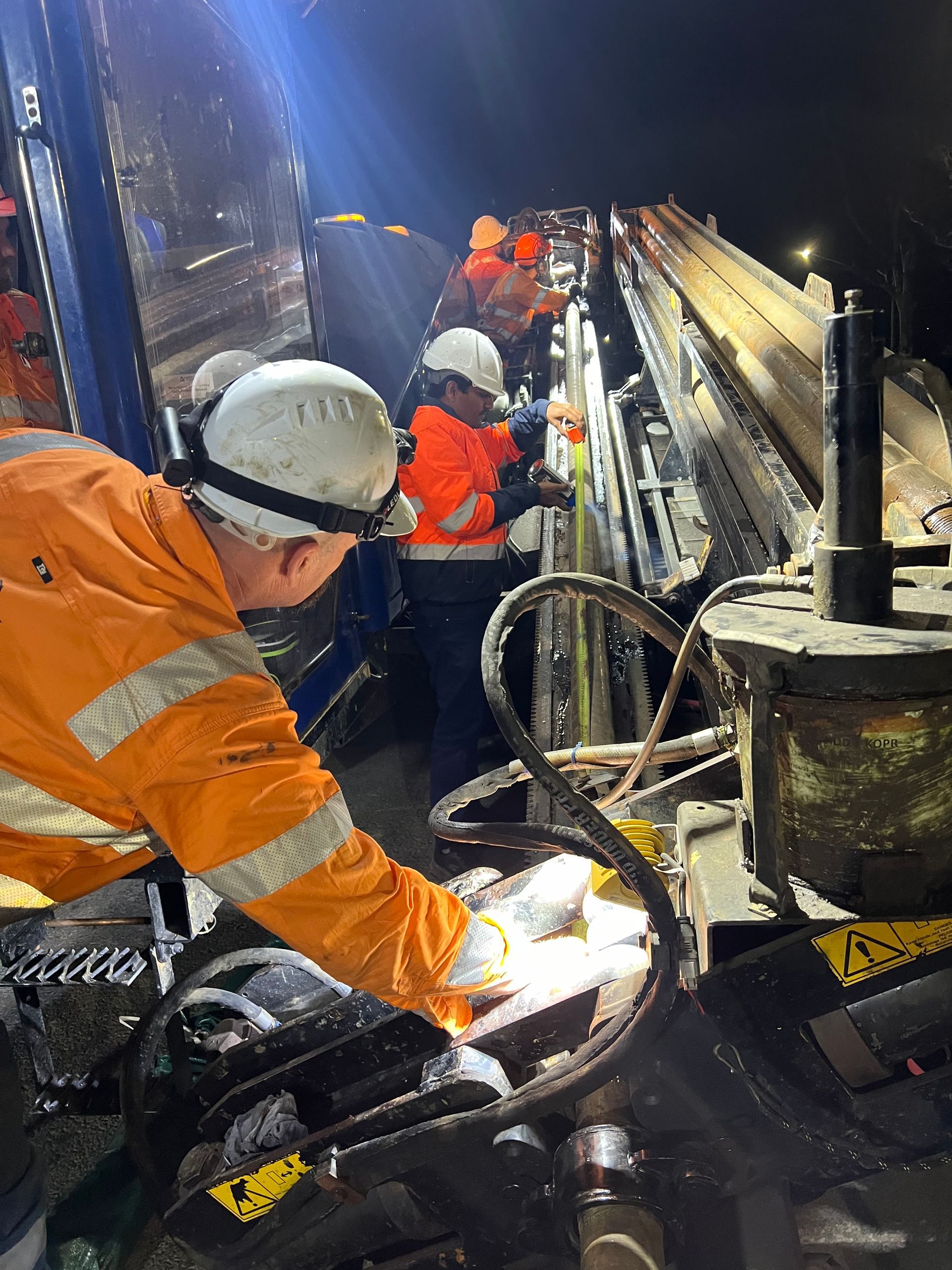 A close-up of a drilling machine on grass with pipes, mud, and the operator partially visible