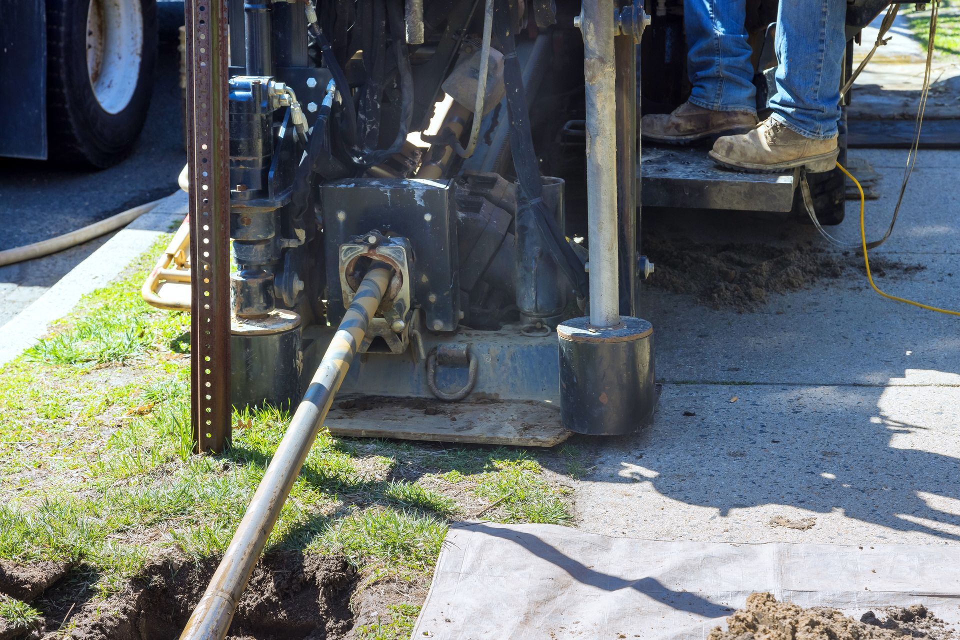 A worker operates a heavy machine to drill an underground pipe on a grassy area near a sidewalk edge