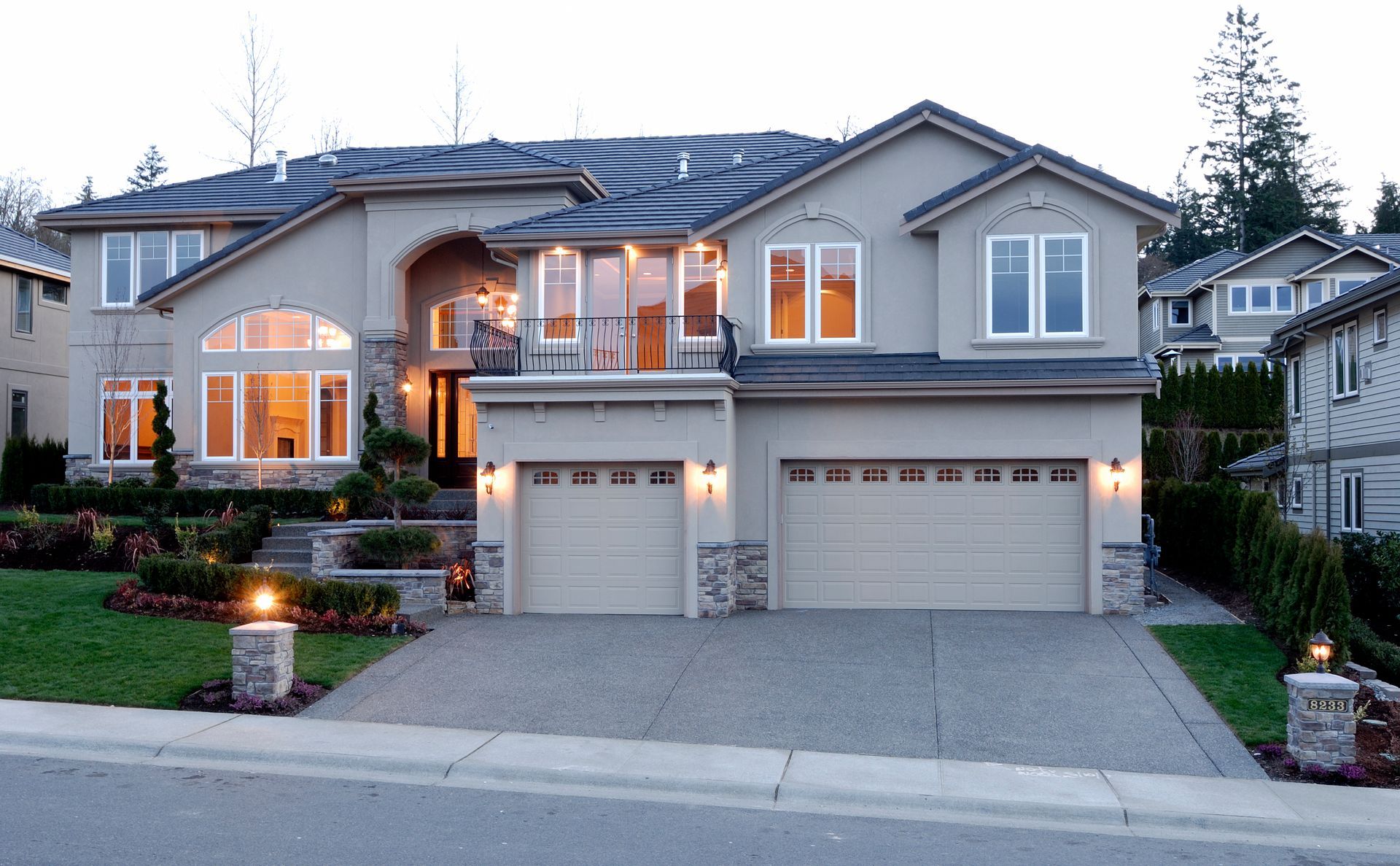 Two-story beige house with a gray roof, two-car garage, and manicured lawn.