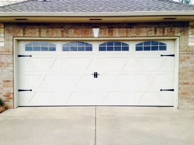 White garage door with arched windows, accented by black hardware, set in a brick and beige building.